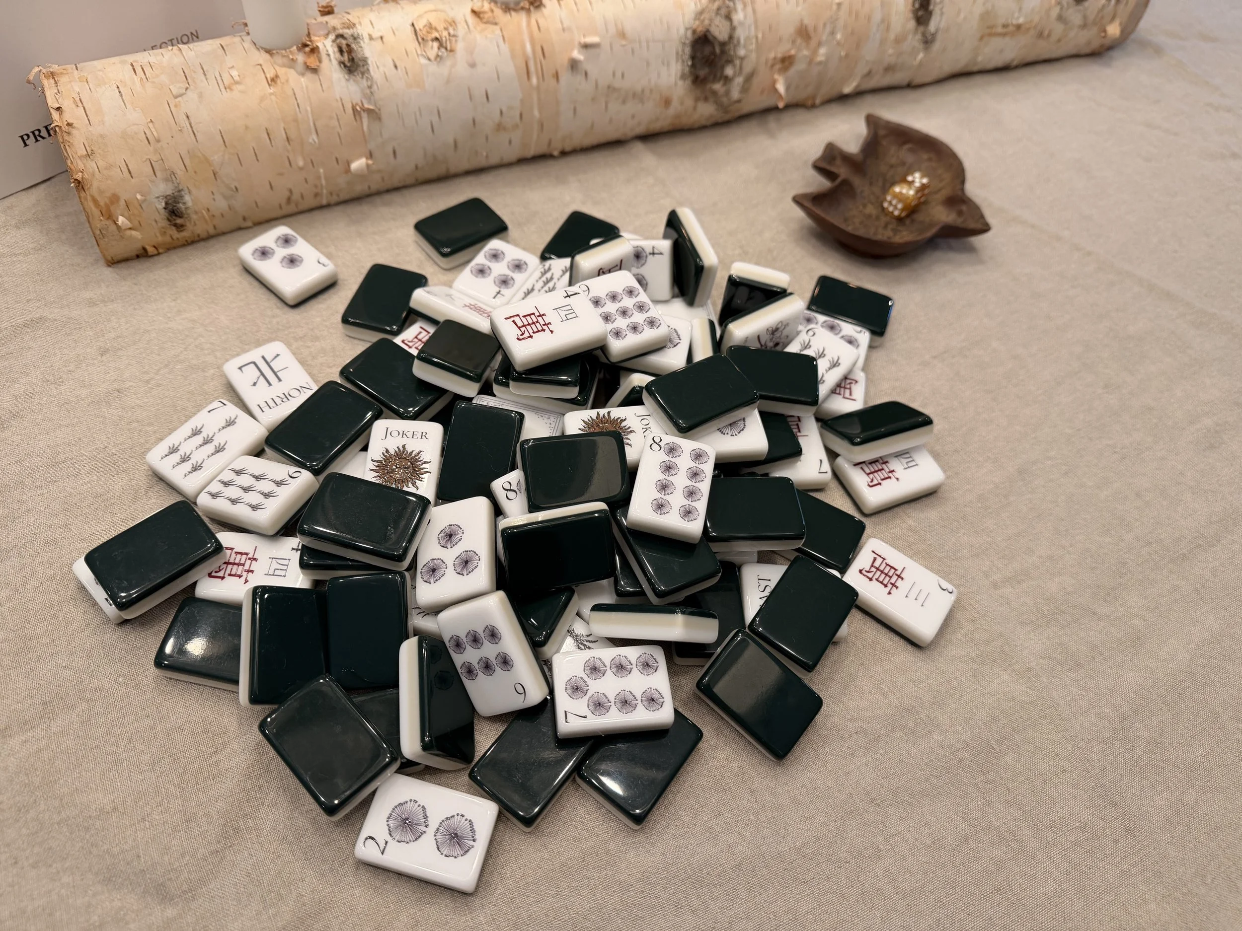 A pile of Winter Solstice tiles, green backs and printed faces mixed, on a linen tablecloth beside a birch log and a small bird-shaped dish holding gold dice.