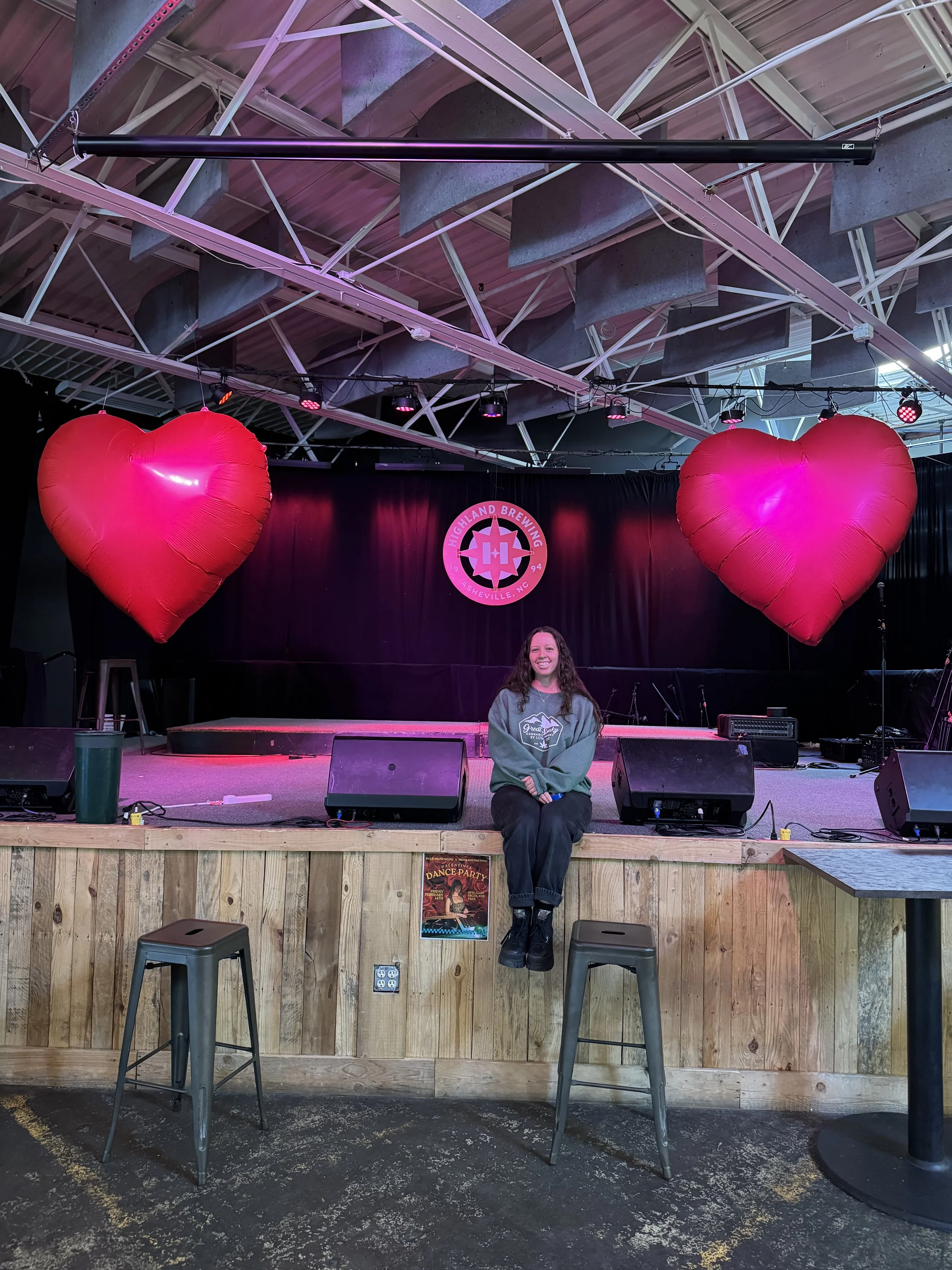A woman sitting on the stage in front of two large pink heart-shaped balloons. Behind her, there is a black curtain with a purple and pink circle logo that says 'Highland Brewing Asheville, NC.' The stage has some microphone equipment and stage lights. The ceiling has exposed metal beams and a ventilation duct.