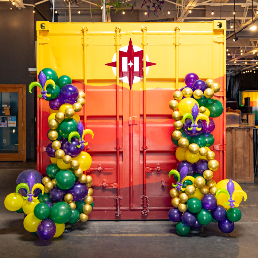Decorative balloon arrangement with purple, green, yellow, and gold balloons forming a vertical column on each side of a bright yellow and orange container backdrop. The balloons feature motifs like a fleur-de-lis and a knight's helmet in Mardi Gras colors.