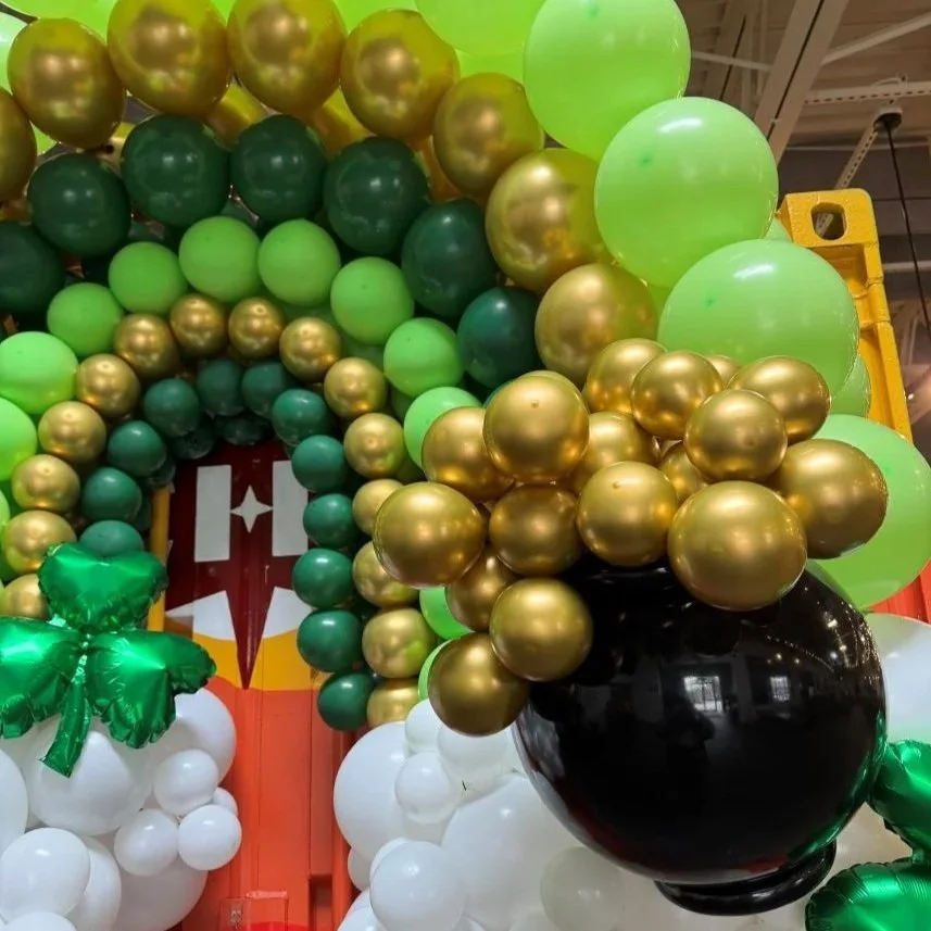 Colorful balloon arch with green, gold, white, and black balloons, featuring a green four-leaf clover decoration, suggesting a St. Patrick's Day celebration.