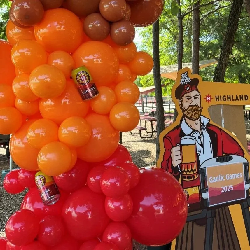 Decorative display of orange, red, and brown balloons next to a cartoon cutout of a man with a beard, wearing a red coat and holding a beer mug, at Highland Gaelic Games 2025 event.