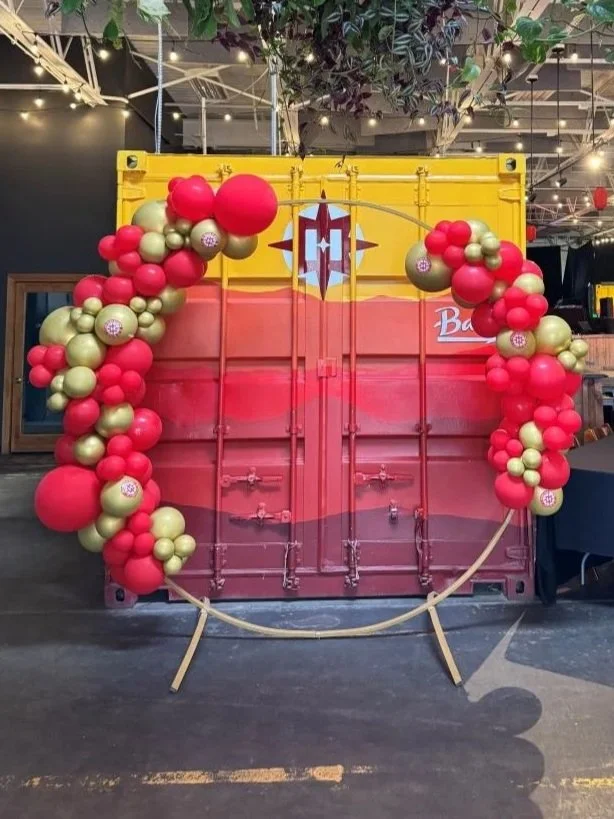 Decorative balloon arch in red and gold colors framing a red backdrop with a heart-shaped design, set against a yellow shipping container. The setup appears to be decoration for a celebration or event, possibly Valentine's Day.