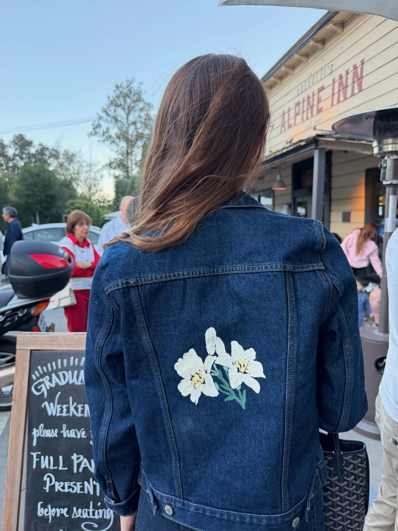 A woman with long brown hair wearing a denim jacket with floral embroidery on the back, standing outside near a restaurant or cafe, with a chalkboard sign nearby and people in the background.
