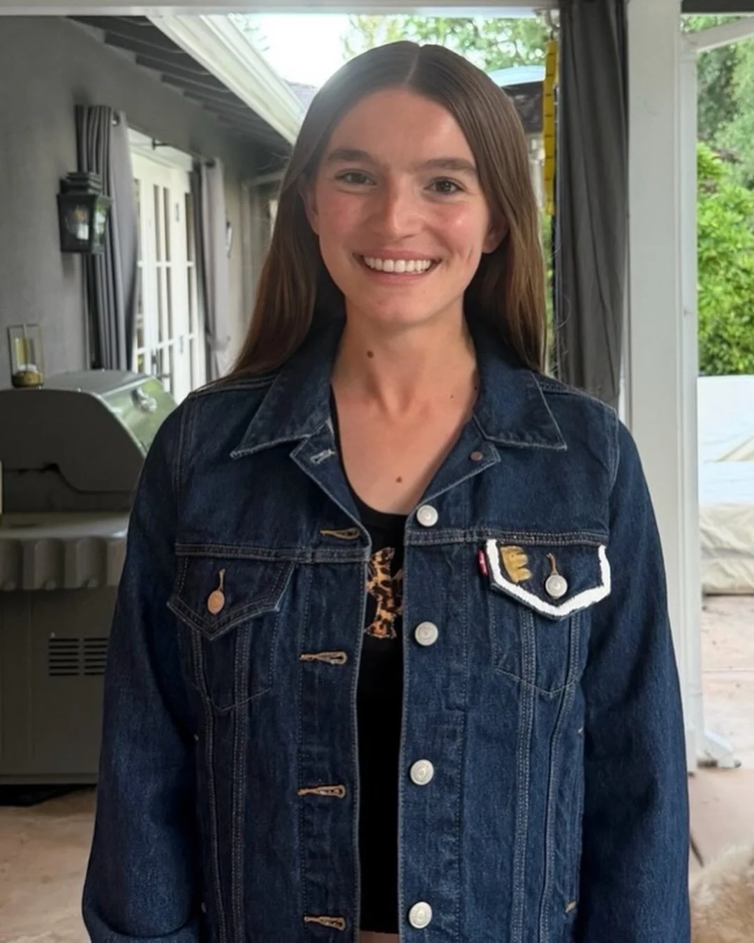 Young woman with long brown hair and a bright smile wearing a denim jacket indoors with greenery and a patio visible behind her.