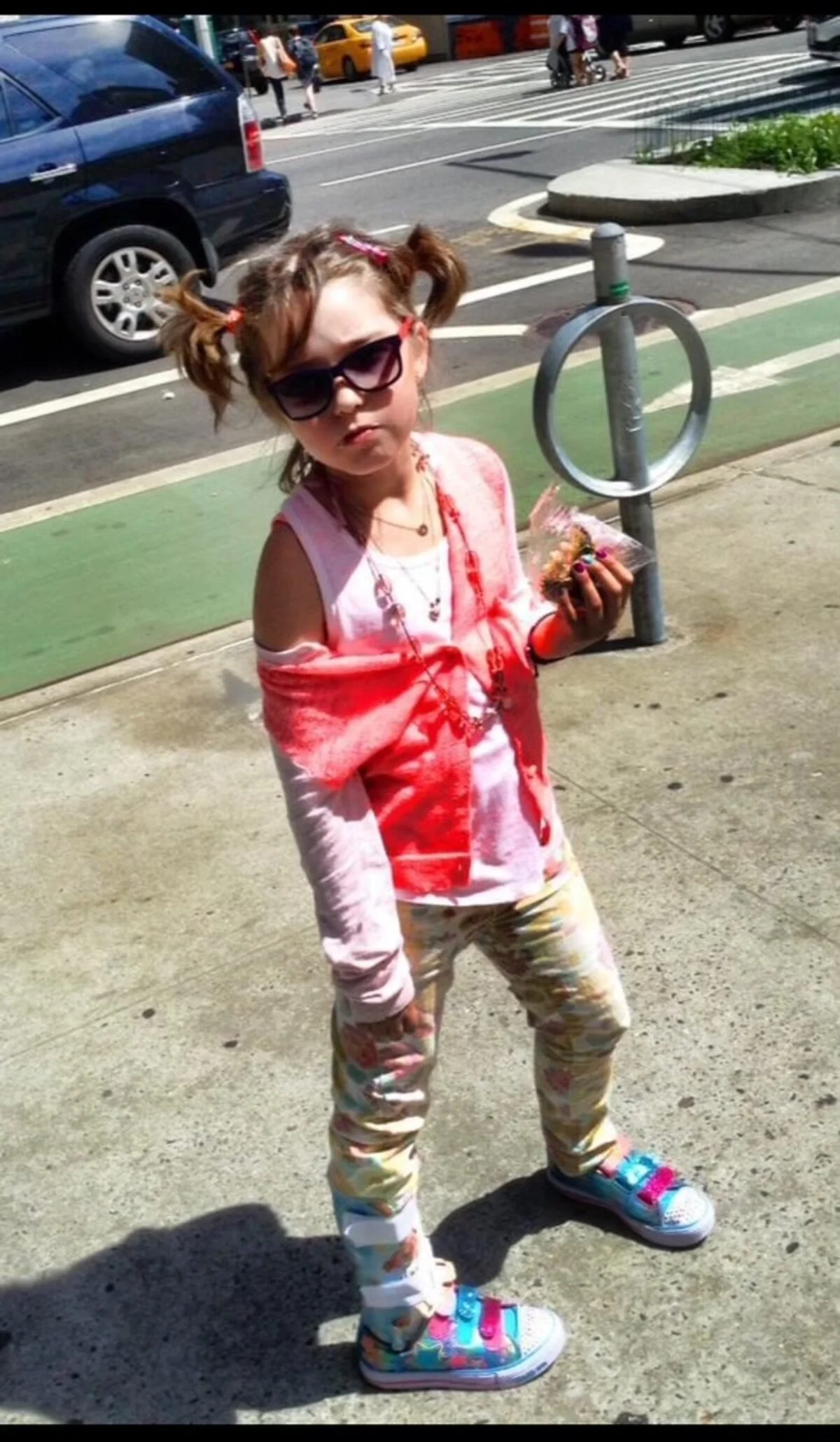 A young Elle, with braided hair, wearing sunglasses, a pink and white tie-dye shirt, matching patterned pants, and colorful sneakers, stands on a sidewalk holding a snack.