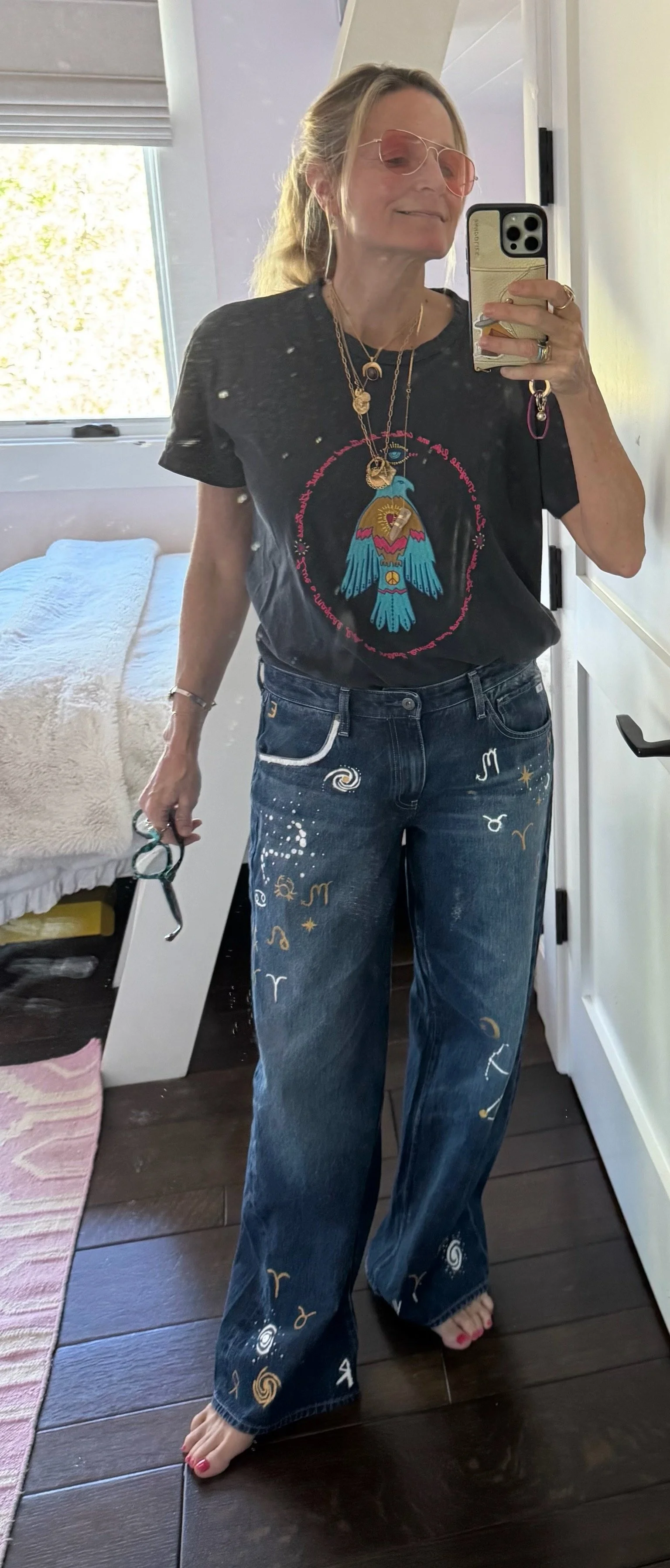 A woman taking a mirror selfie in a bedroom. She is wearing pink sunglasses, a black horoscope-themed t-shirt with a blue bird graphic, and wide-leg jeans decorated with zodiac symbols and spirals. She has layered necklaces, rings, and is holding a p