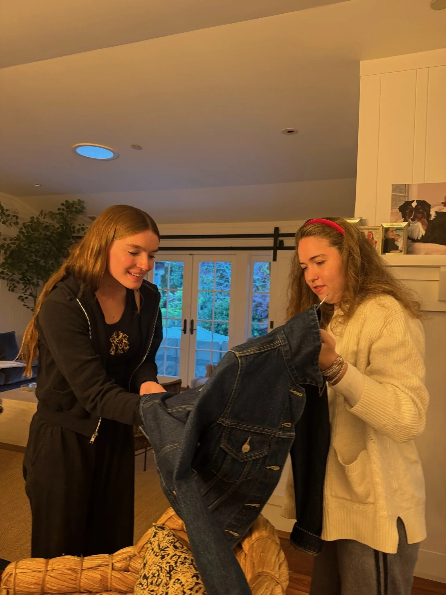 Two young women are smiling and interacting as they look at a pair of jeans hanging on a clothing rack inside a cozy, well-lit room. The room has large glass doors leading outside, a potted plant in the corner, and family photos on the wall above the