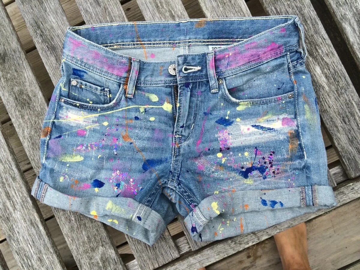Pair of denim shorts with colorful paint splatters, lying on a wooden surface.