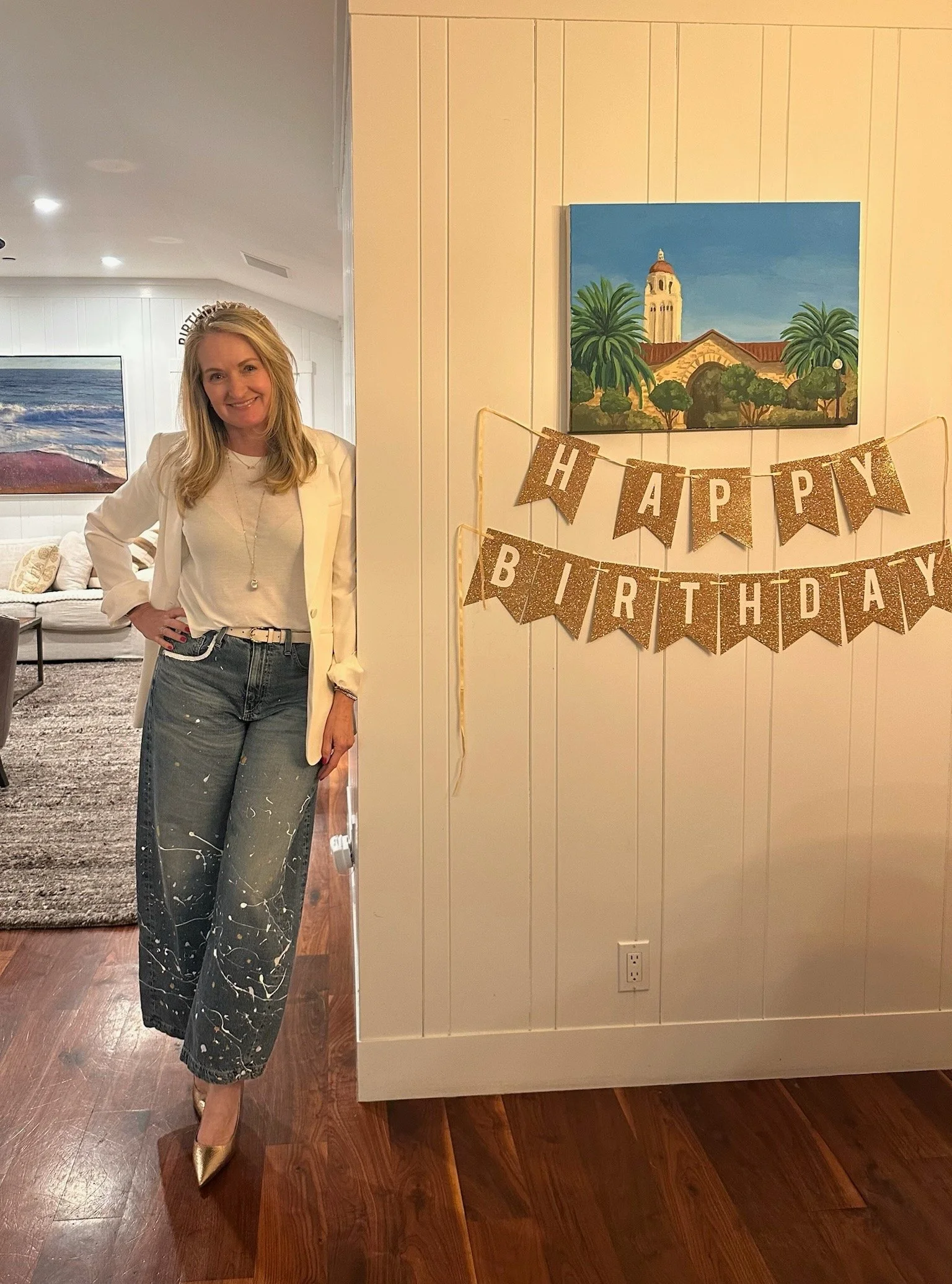 A woman dressed in a white blazer, a white top, and gray jeans with splatter paint design, standing indoors with a smile. There are birthday decorations on the wall that say "HAPPY BIRTHDAY" in gold glitter letters. A colorful painting of a building 