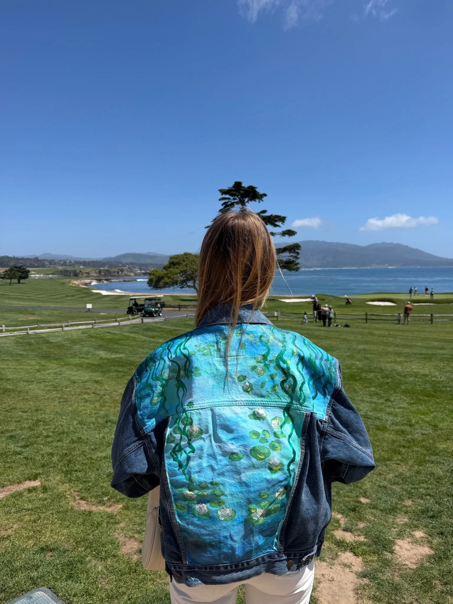 A person with long hair wearing a denim jacket with a colorful, abstract pattern on the back, standing on a golf course and overlooking a body of water with hills in the background.
