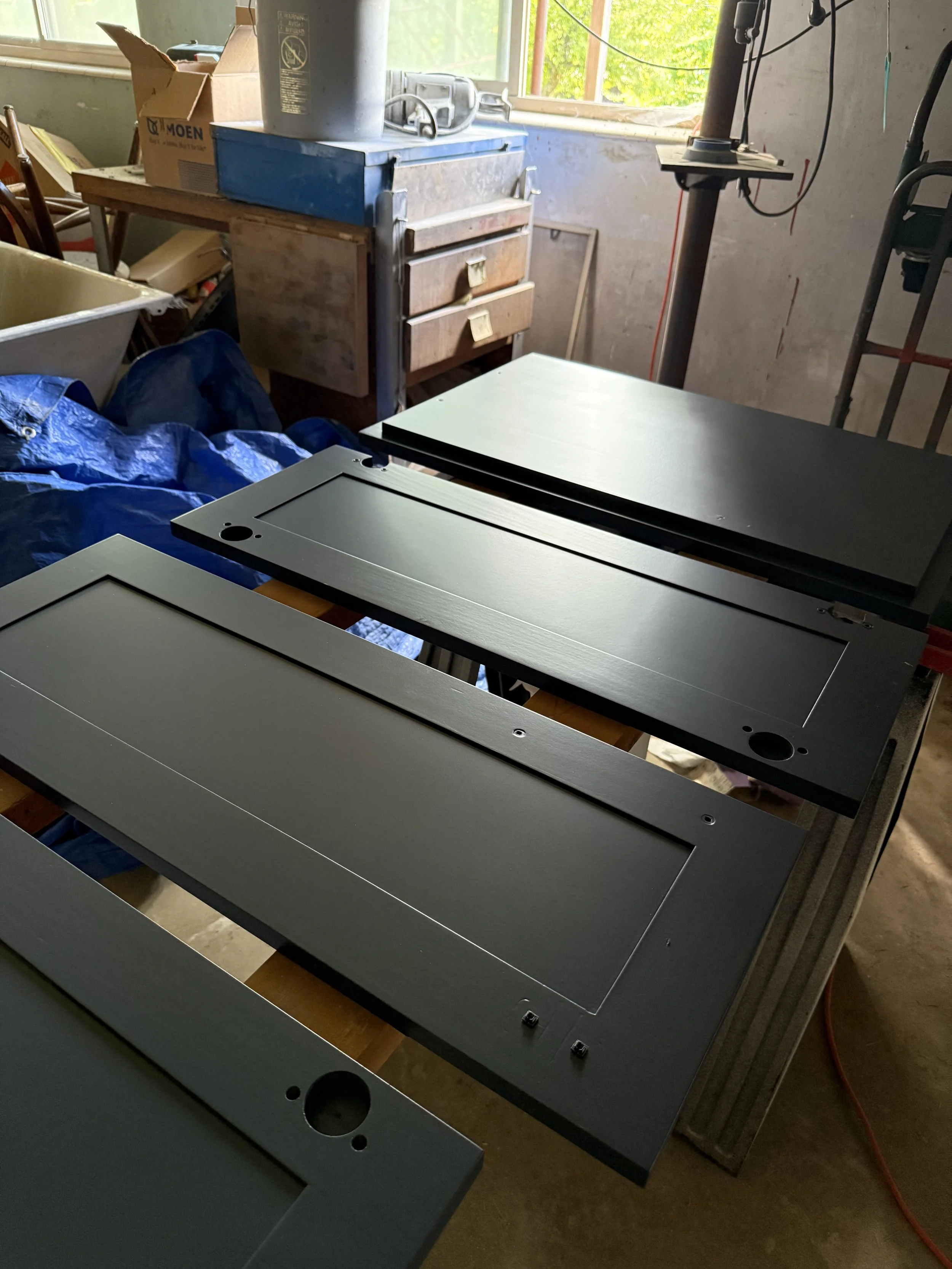 An indoor workshop or storage room with two black metal cabinets or drawers on a wooden table, with a window showing greenery outside in the background, and various tools and boxes scattered around.