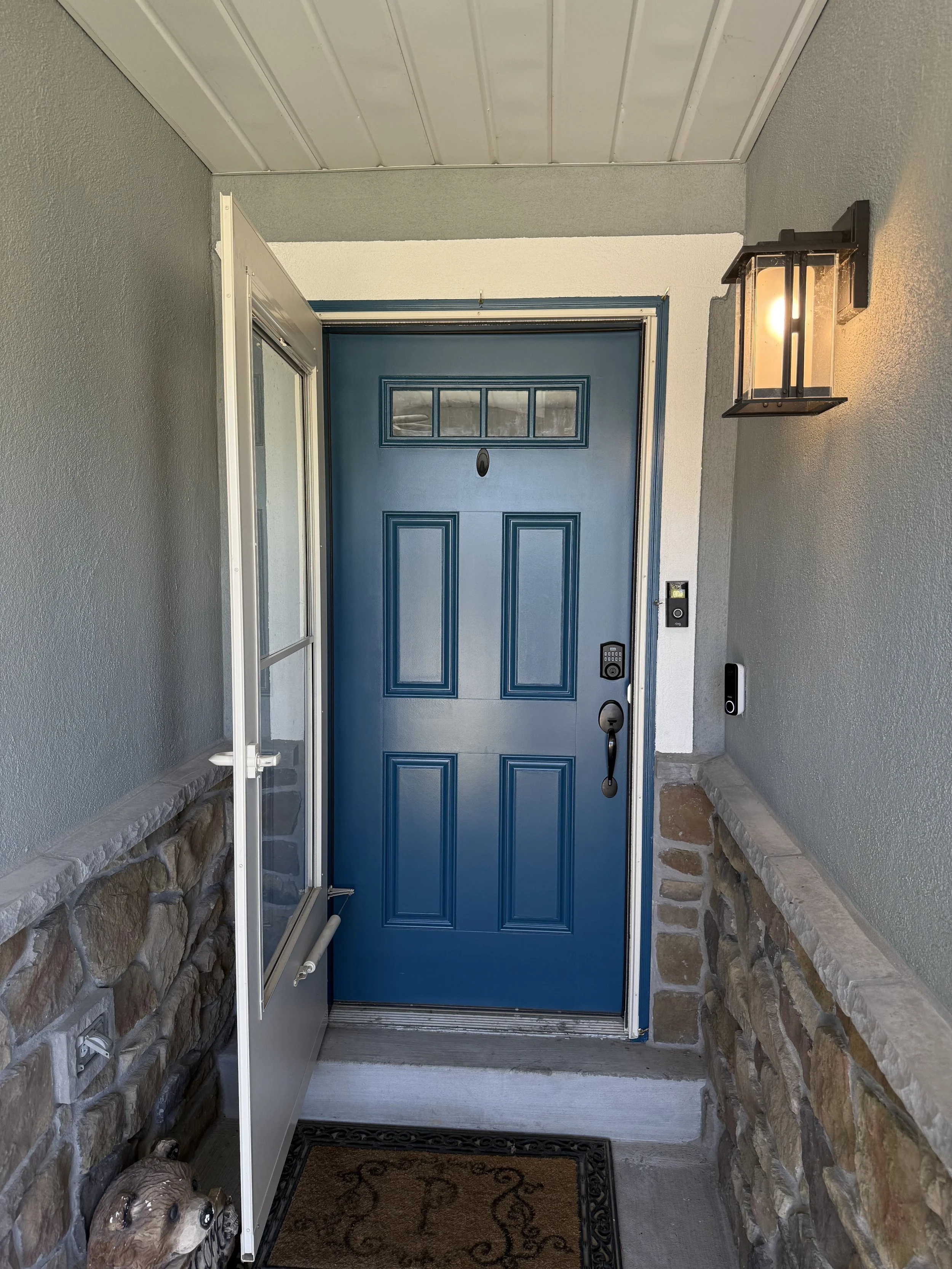 Front porch with a blue door, stone walls, a light fixture, and a doormat.