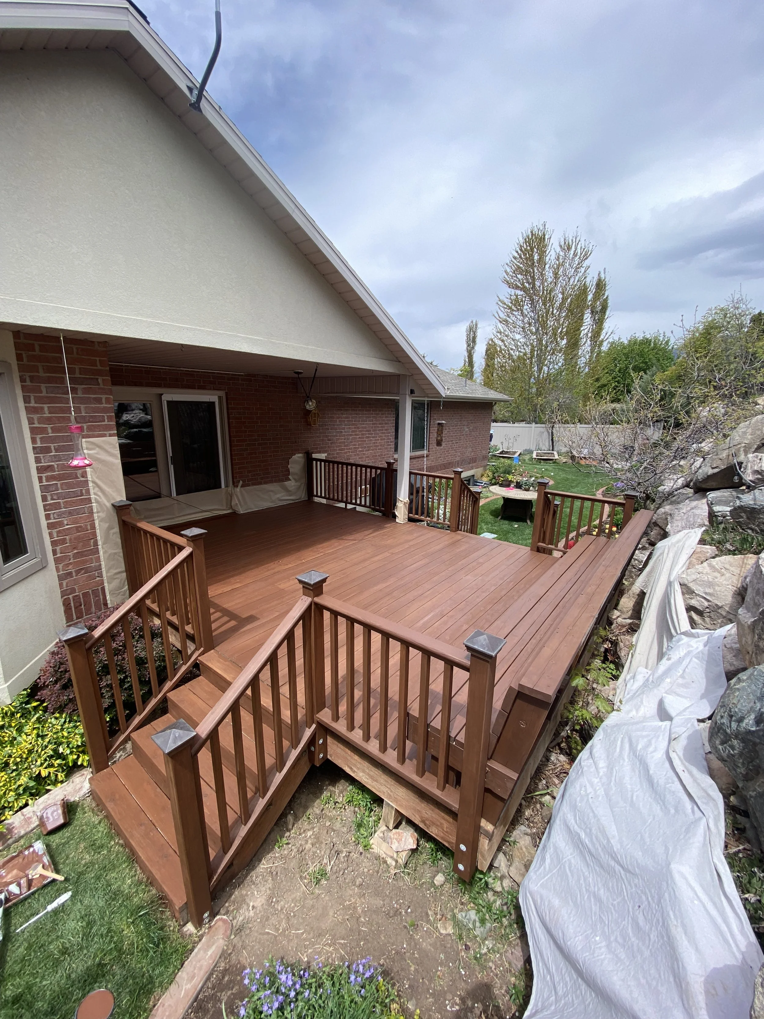 A newly built wooden deck attached to a house with a brick exterior. The deck has stairs and wooden railings. Surrounding the deck are plants, flowers, and garden decorations. There are trees and a fence in the background under a partly cloudy sky.