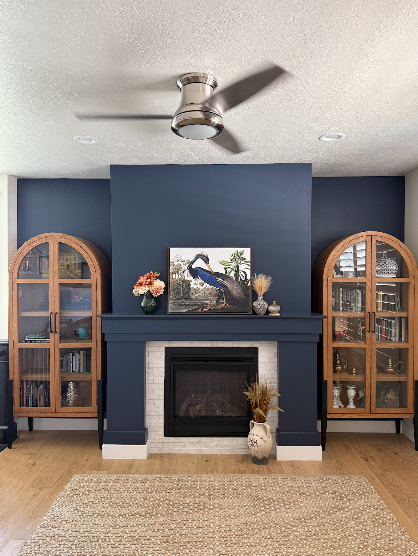 Living room fireplace with navy blue mantel, two glass-front wooden cabinets, artwork of a heron, vases, and decorative items.