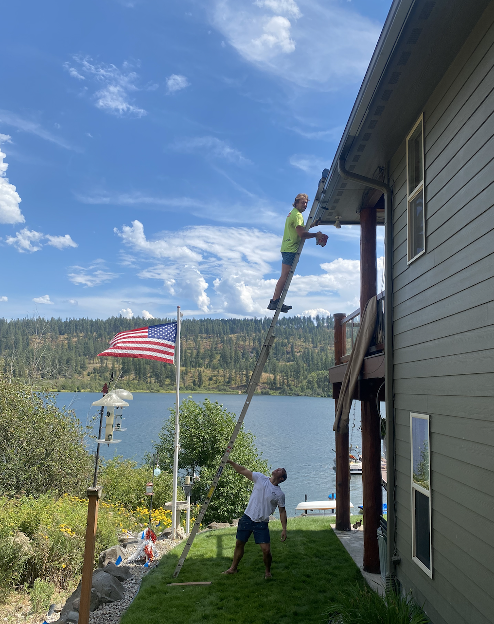 Two men repair the gutter of a house by a large lake with a forested hill in the background. One man stands on a ladder leaning against the house, holding a tool, while the other man supports the ladder from below. The house is beige with siding, and there is an American flag on a pole near a bird feeder, with colorful flowers and landscaping around.