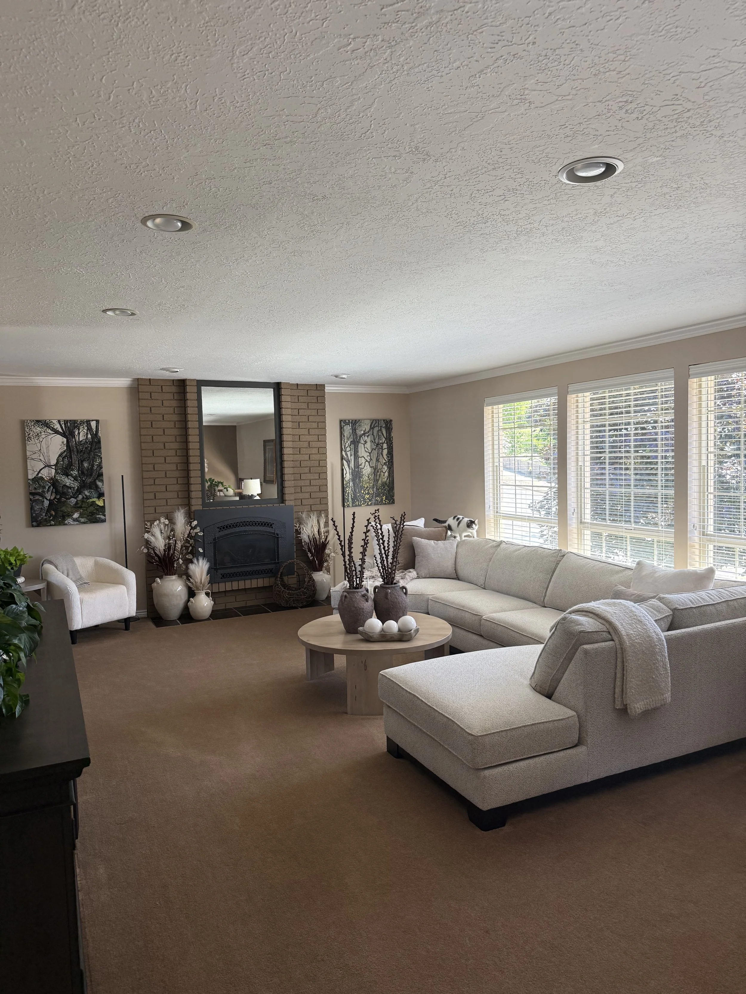 Living room with beige carpet, white sectional sofa, decorated with throw blankets and pillows, large windows with blinds, brick fireplace with mirror and artwork, open doorway with visible furniture, potted plants, and a cat sitting on the window si