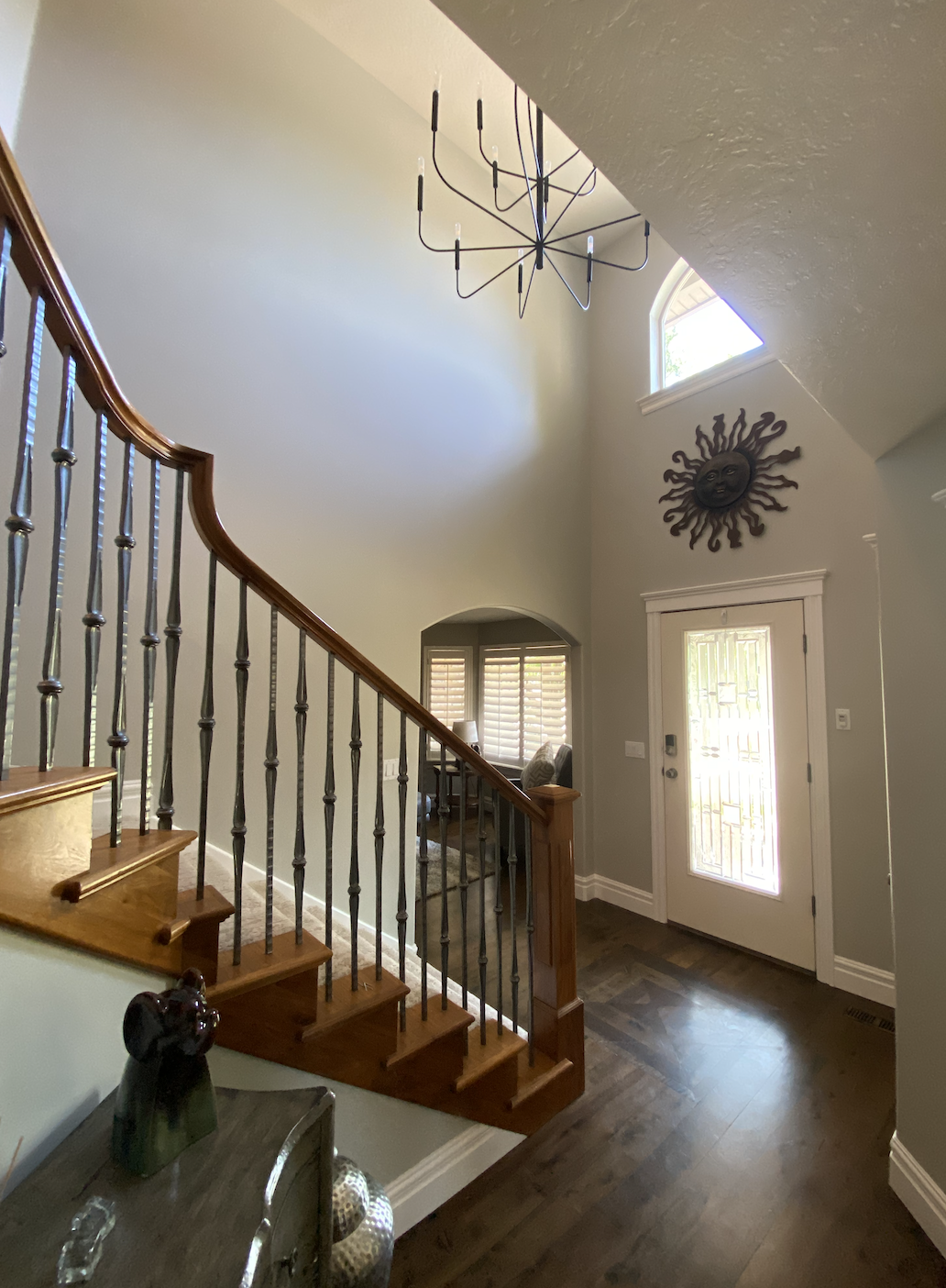 Entryway with a white front door, small window above, sun face wall decor, staircase with wooden handrail and metal balusters, dark wood flooring, and a small seating area with window shutters.