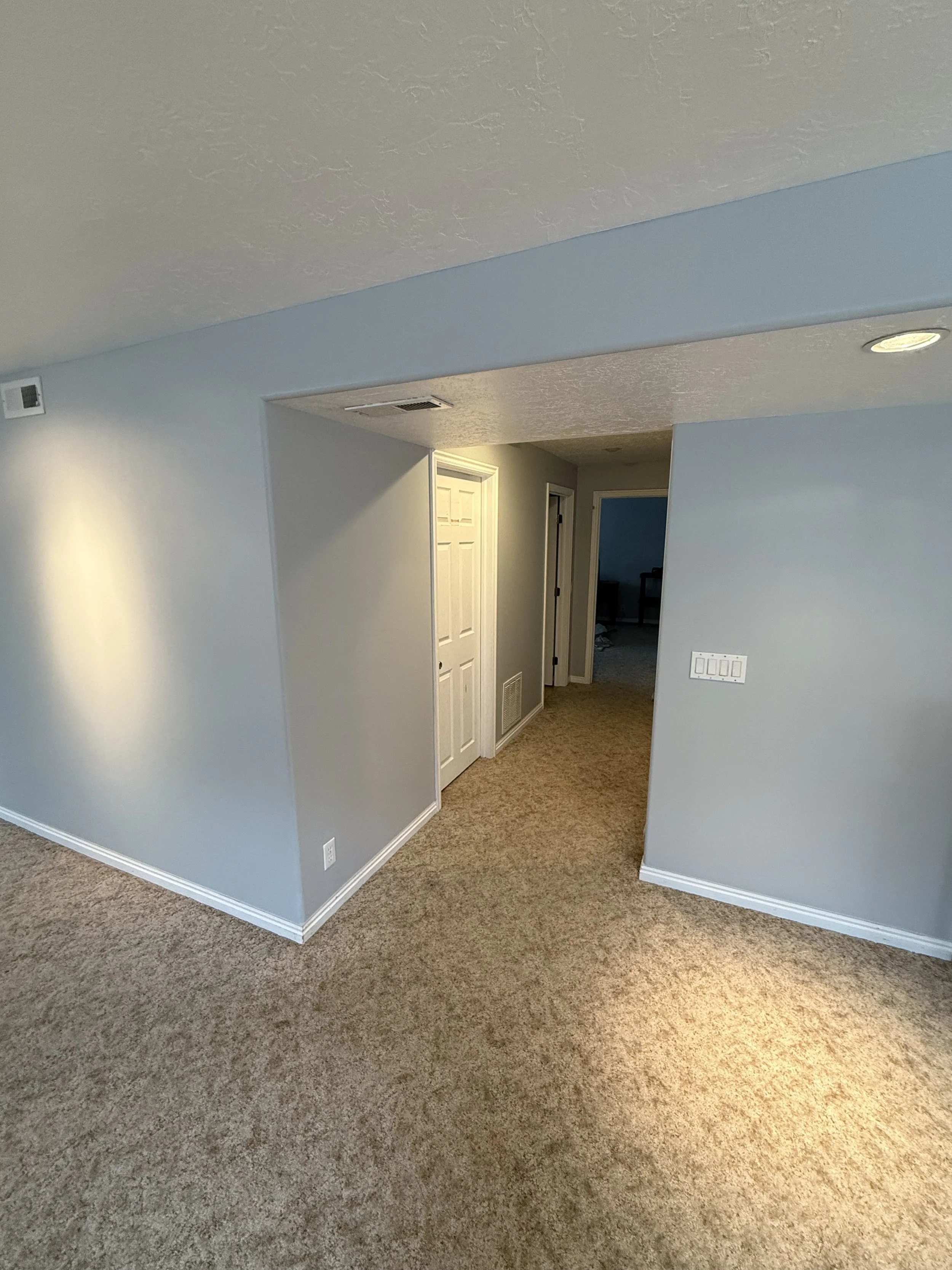 Interior view of a residential house with beige carpeted floors, light blue walls, and a doorway leading to a room with a dark piece of furniture.