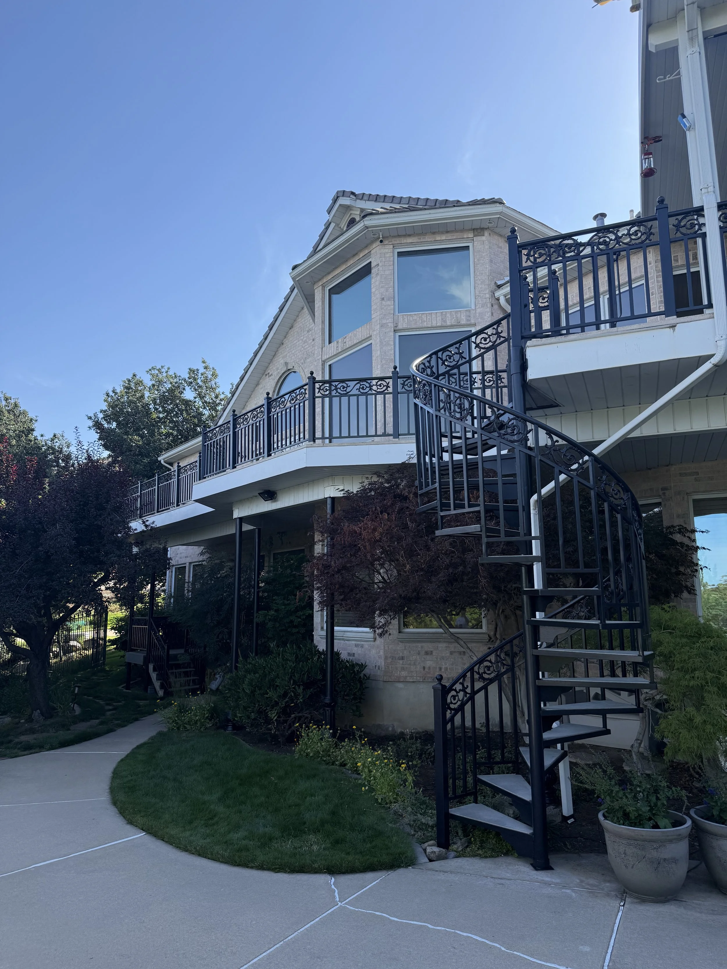 A multi-story house with a large windowed front, a spiral staircase railing, and a backyard garden with trees and a sidewalk.