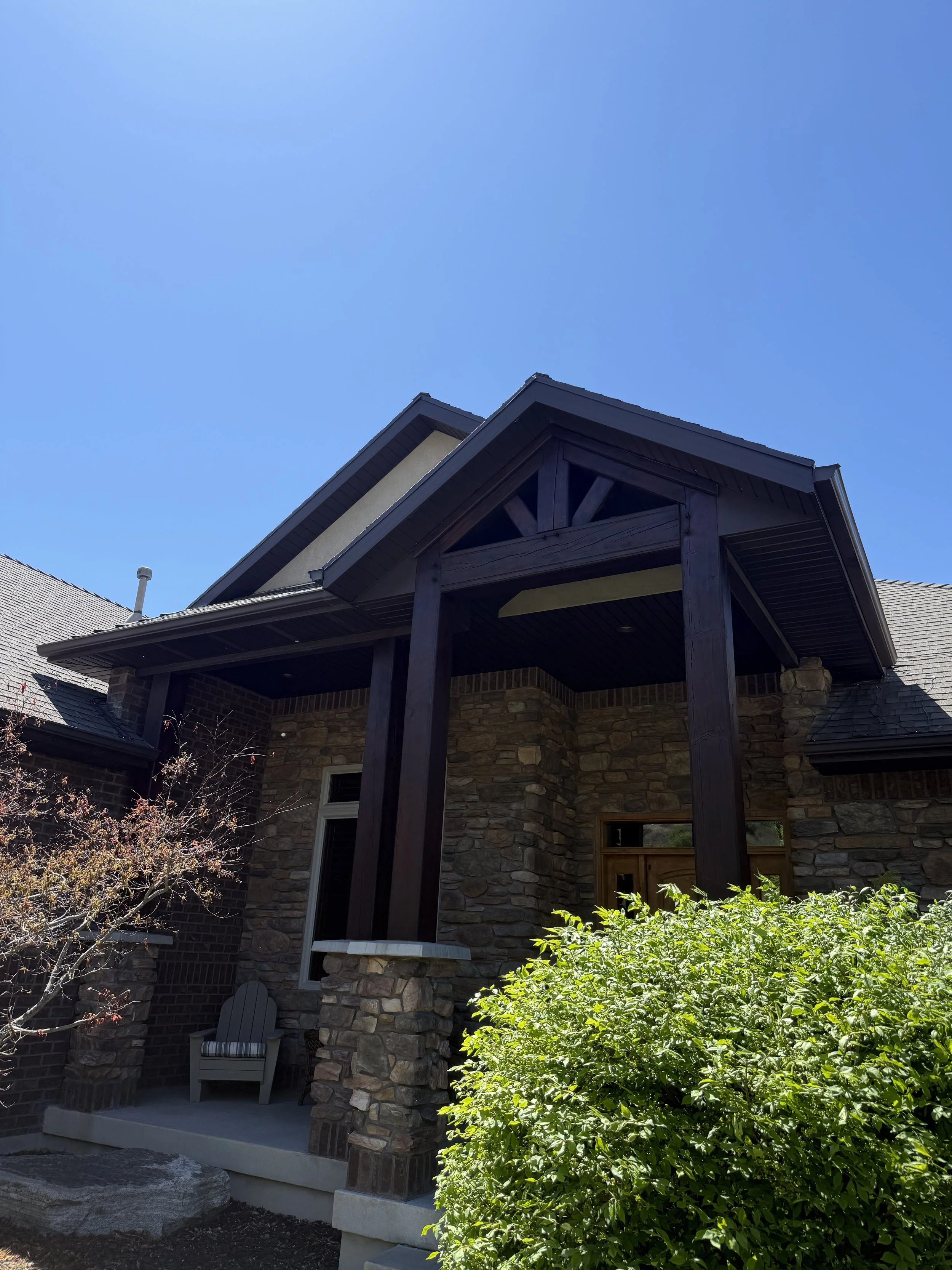 View of a house with stone and brick exterior, a covered porch with dark wooden beams, a white lawn chair, and greenery in the foreground under a clear blue sky.