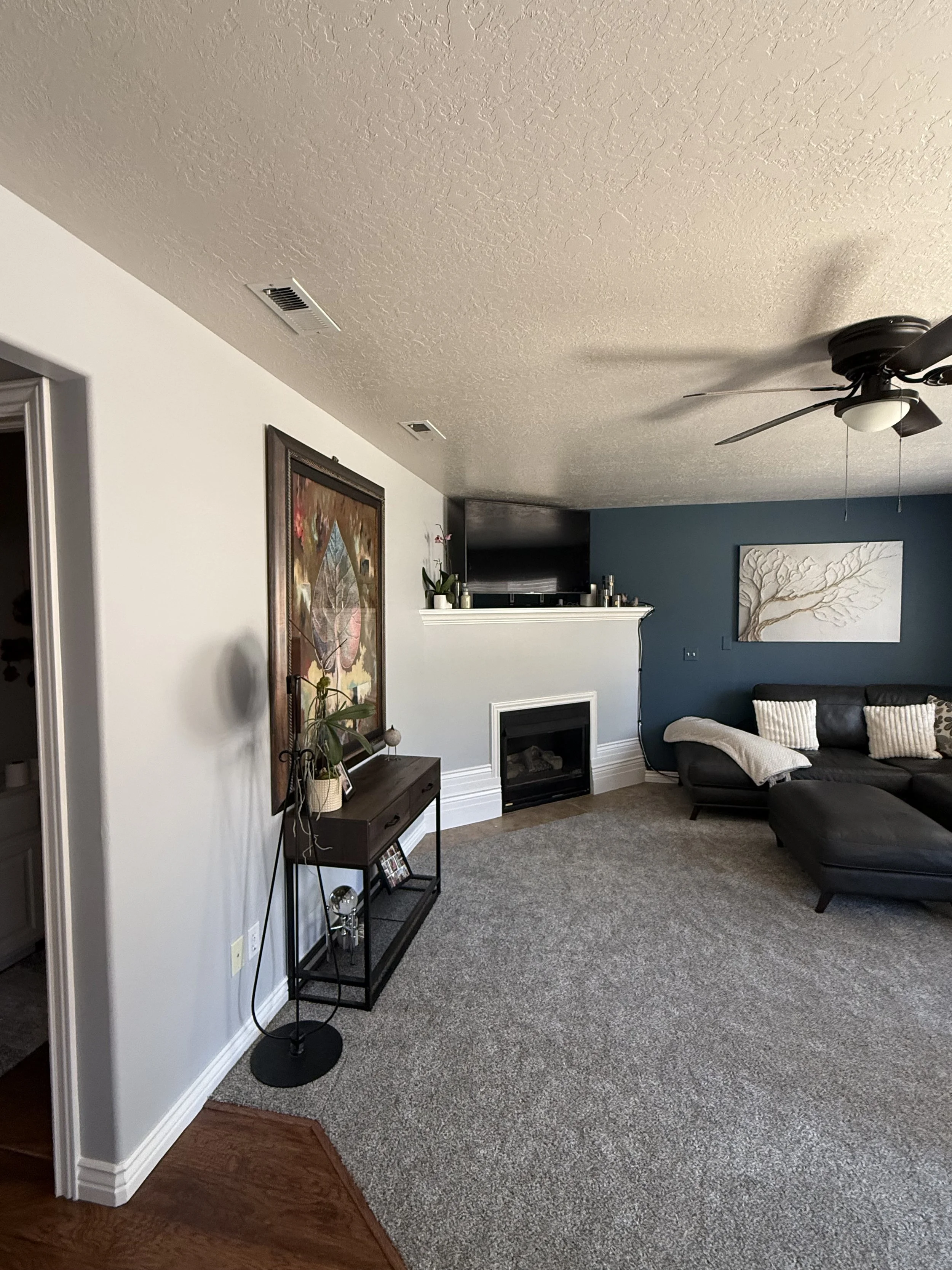 Living room with gray carpet, a dark gray sectional sofa with beige and patterned pillows, a white framed painting of a tree on a blue wall, a black metal and wood console table with decorative items, a white fireplace, a ceiling fan, and a white pop