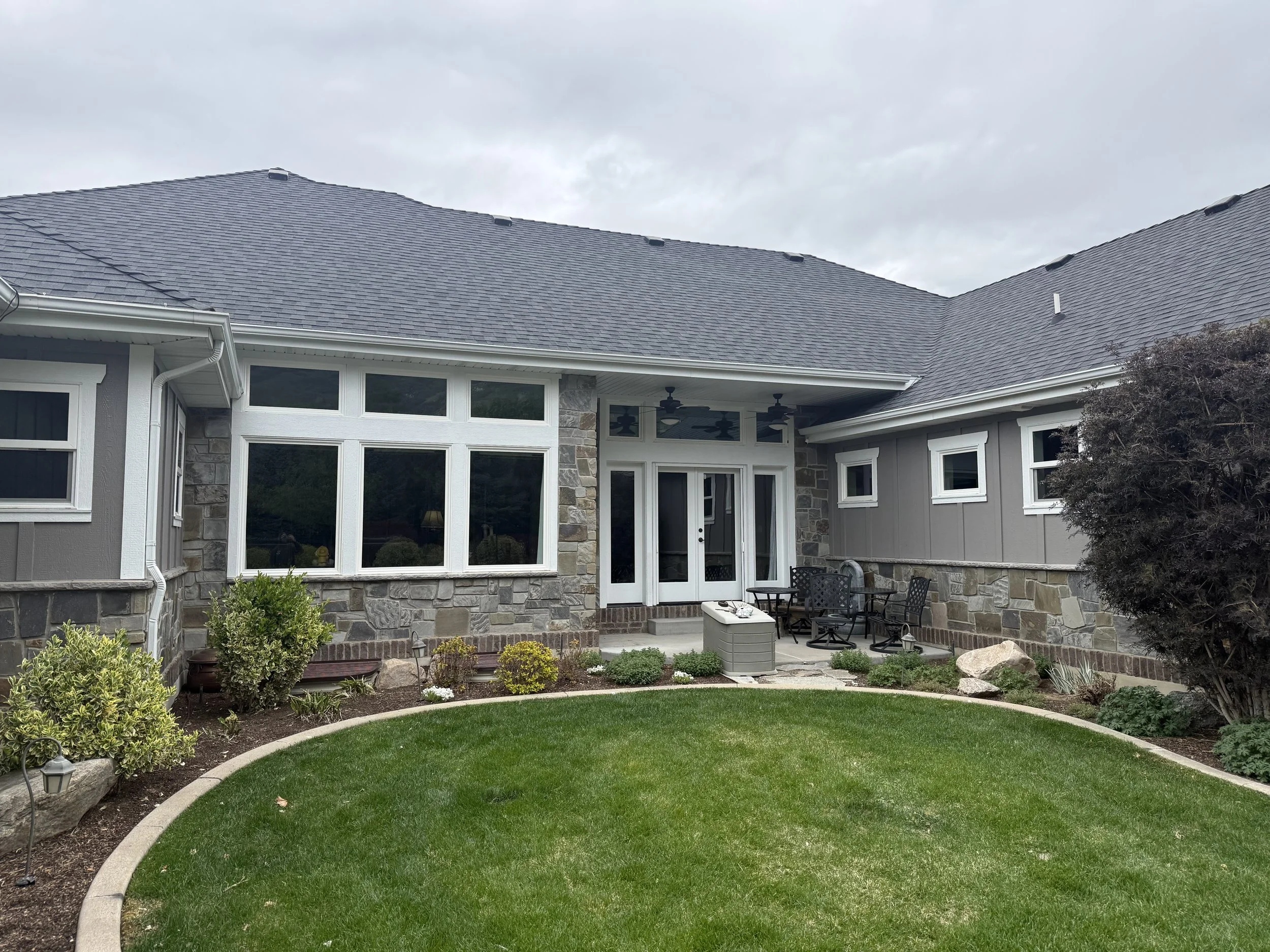 Backyard with green lawn, stone and siding house exterior, patio furniture, garden bed, shrub, and overcast sky.