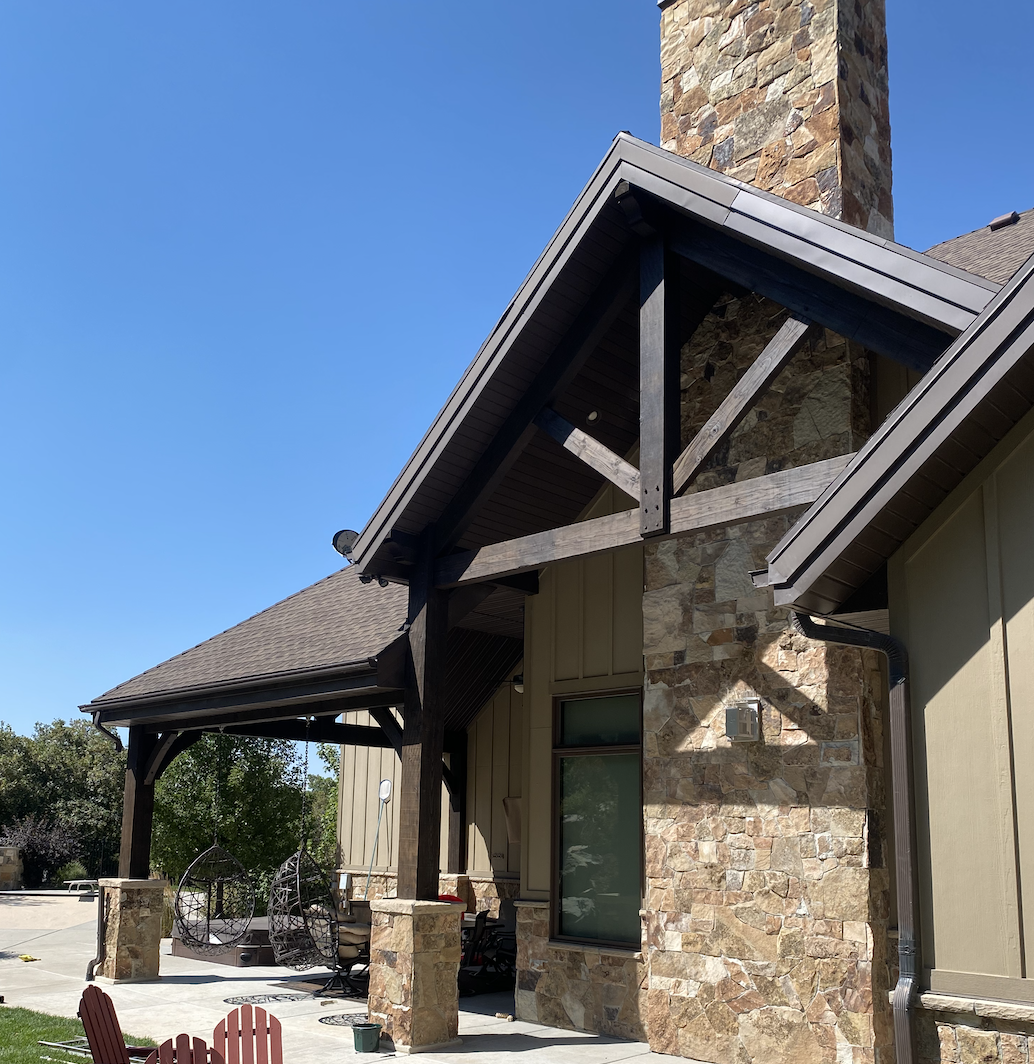 Side view of a modern house with a stone chimney, wooden beams, and a covered patio with outdoor furniture and hanging chairs.