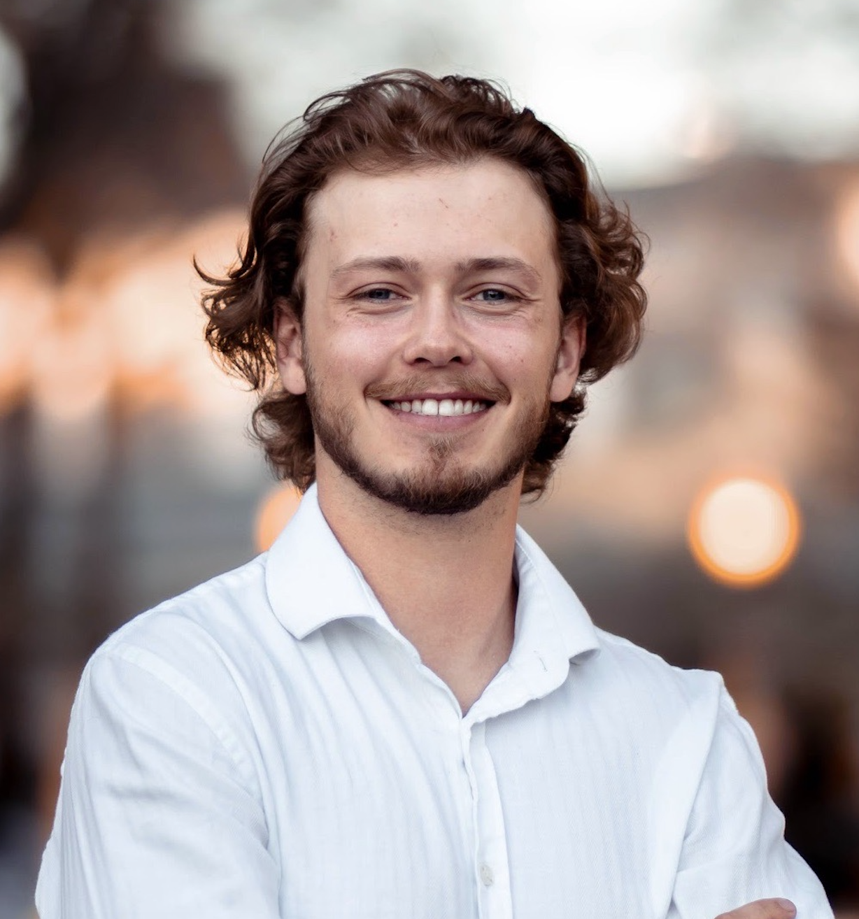 A young man with curly brown hair, a beard, smiling, wearing a white shirt, outdoors with blurred lights in the background.