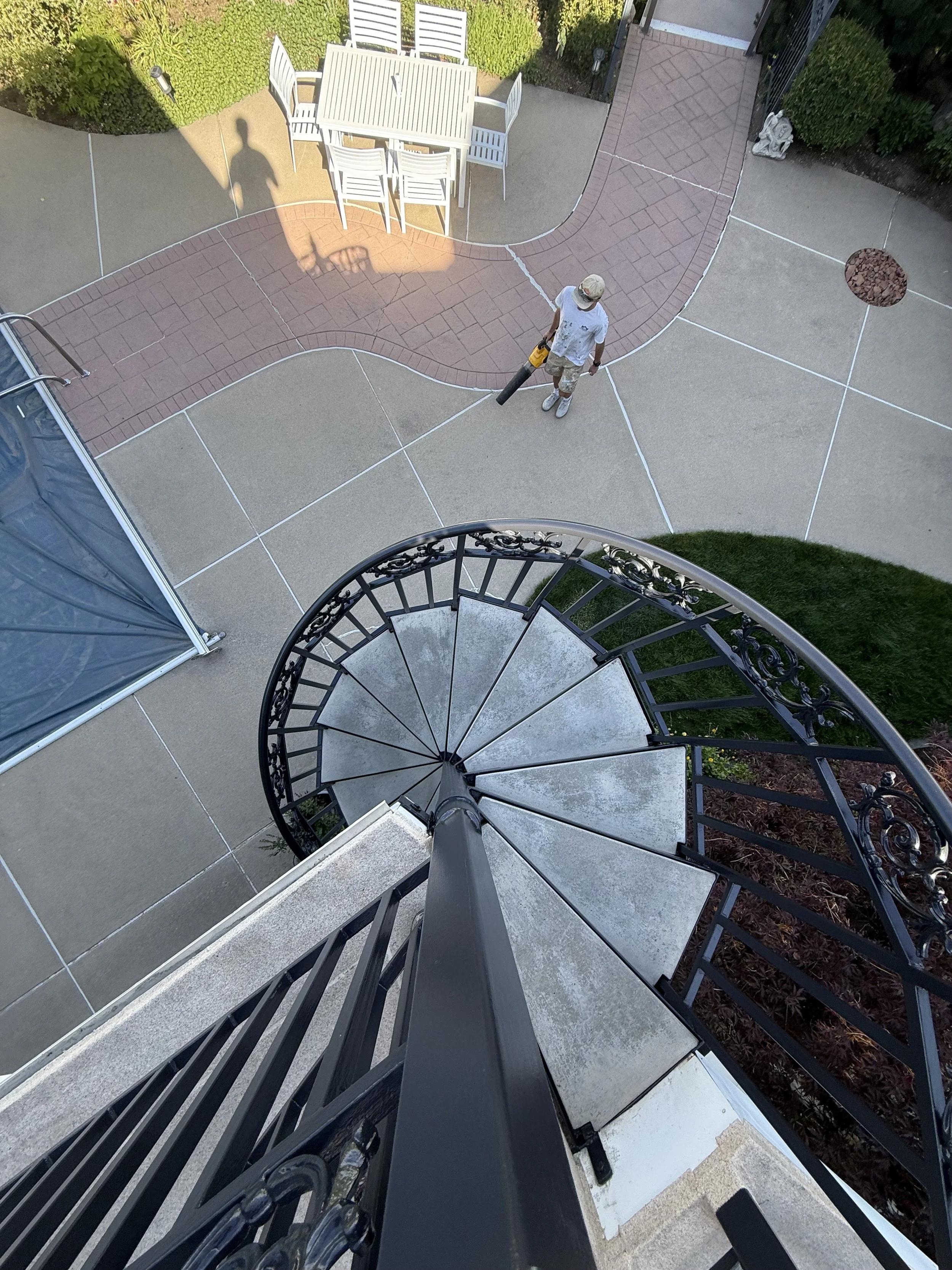 A person in a white t-shirt, camouflage shorts, white shoes, and a cap is cleaning the patio with a leaf blower. The photo is taken from the top of a spiral staircase looking down at the person on the patio with white outdoor furniture and garden pla