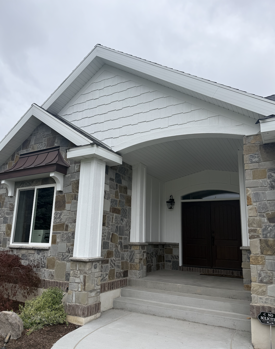 Front view of a modern house with stone exterior, white siding, and a dark wooden door, under an overcast sky.