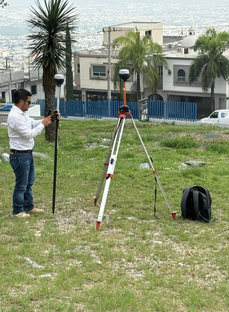Hombre configurando un equipo de medición con un trípode en un área con césped y palmeras, con casas y colinas de fondo.