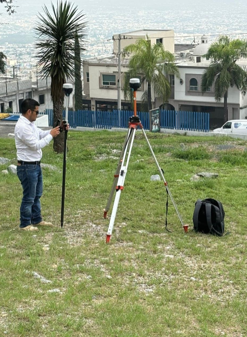 Hombre ajustando un equipo de topografía con un tripié, en un área de césped con casas y palmeras en el fondo, y vista al mar.
