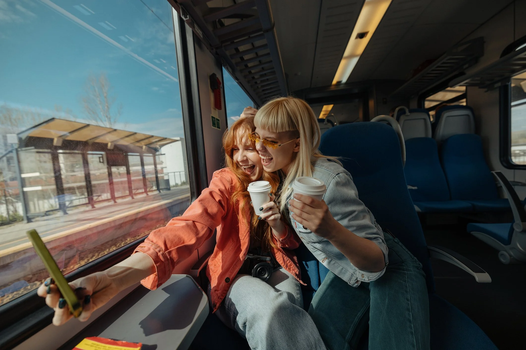 Two young women sitting on a bus, taking a selfie together and smiling, with the woman on the left holding the phone in her hand. They are holding coffee cups, and the bus window shows a cloudy sky and a train station platform outside.