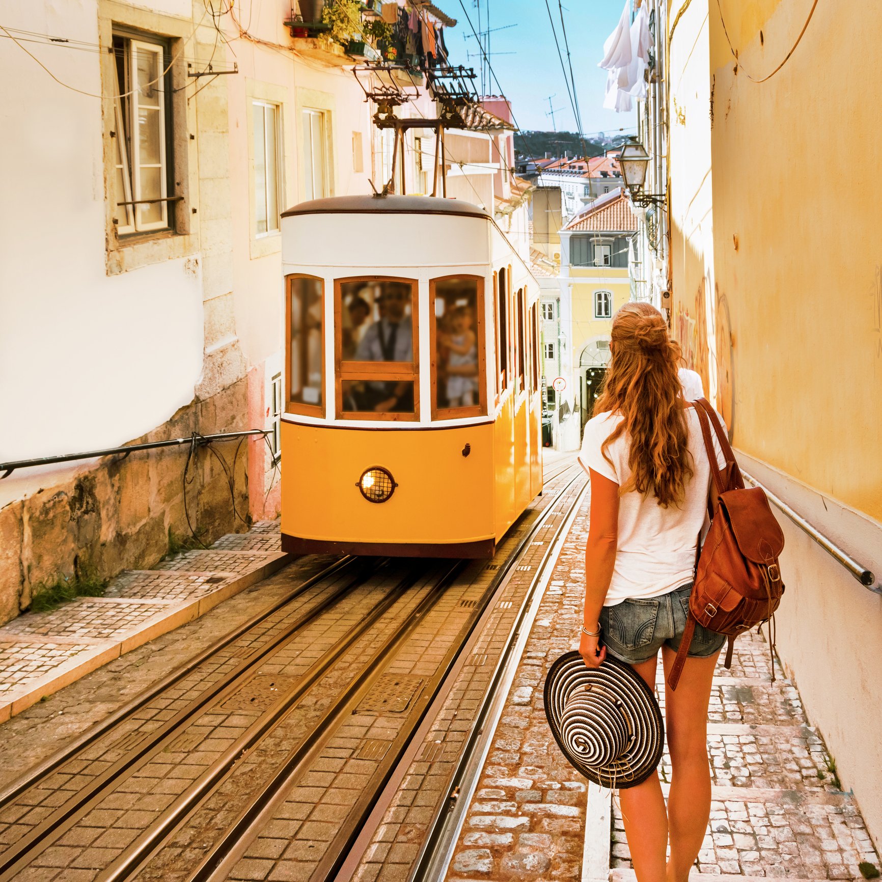 A woman with long hair, wearing a white t-shirt and denim shorts, carrying a brown bag on her shoulder, walking along a street with yellow and white buildings. She holds a black and white striped hat in her left hand. There is a yellow tram on tracks in front of her, and laundry hanging on clotheslines above.
