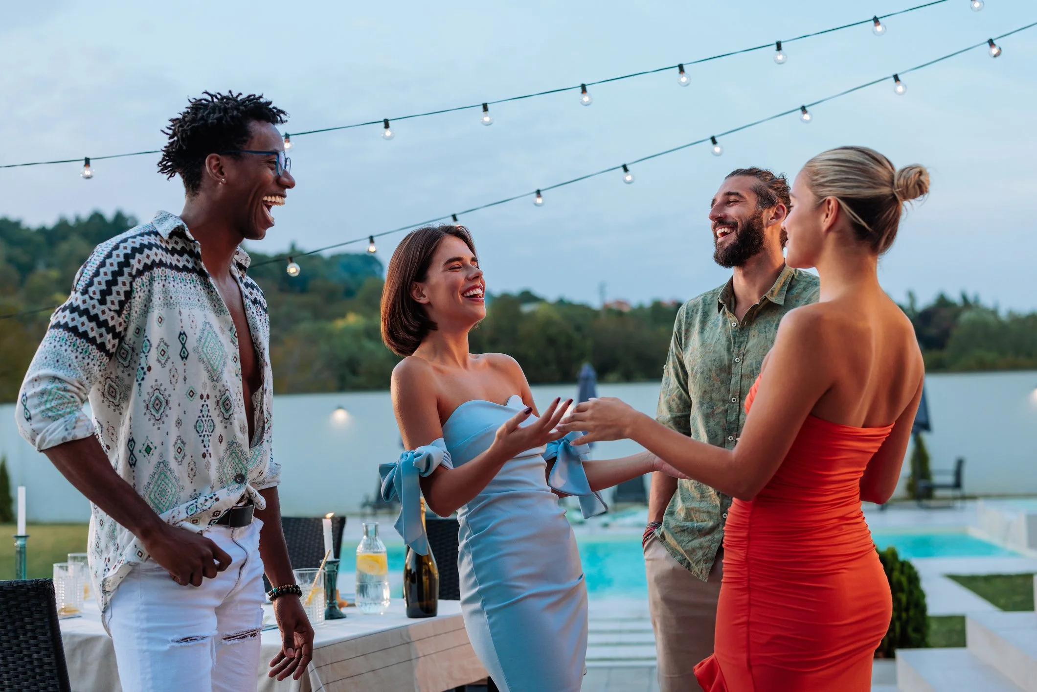 Group of four people enjoying an outdoor gathering, smiling and talking, at sunset with string lights overhead, near a swimming pool, with trees in the background.