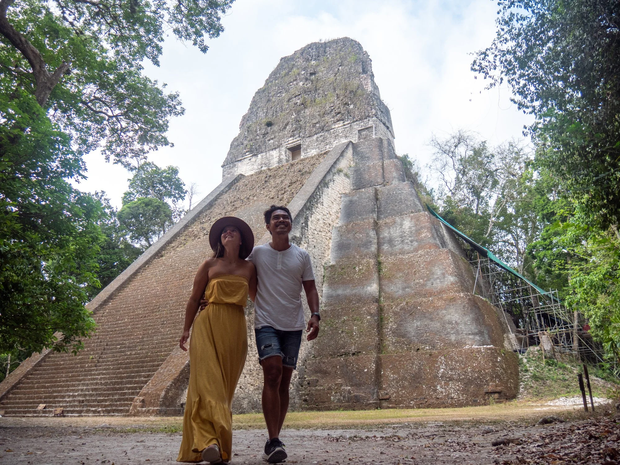 Two tourists, a woman in a yellow dress and a man in a white shirt and denim shorts, walk down stairs in a jungle near an ancient stone pyramid.