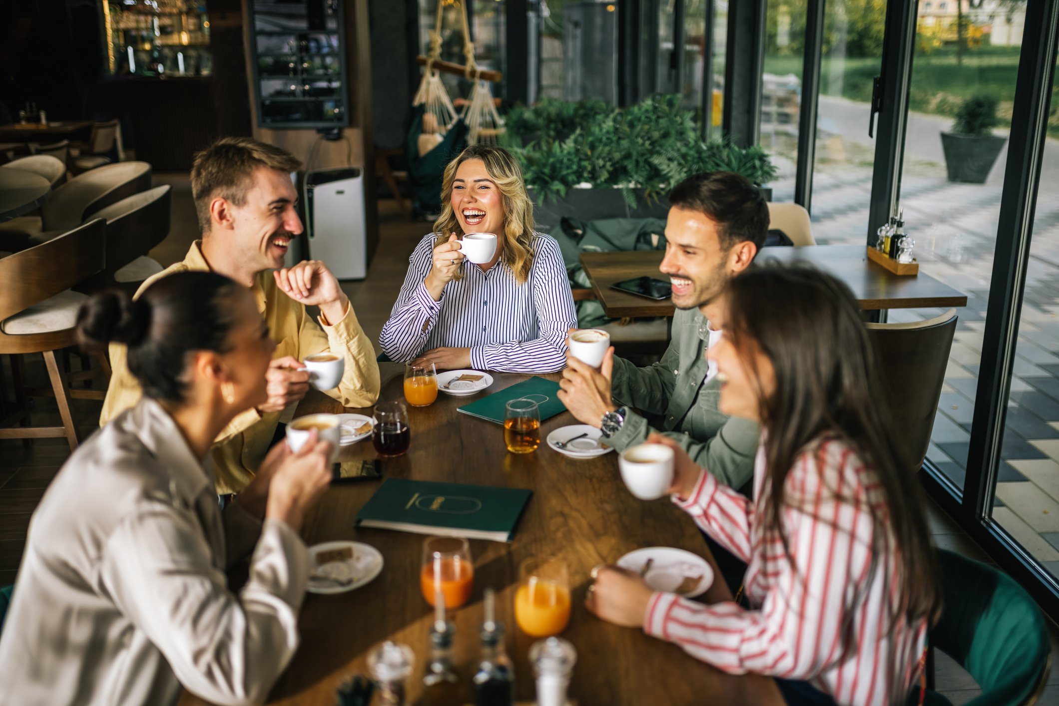 Group of six young adults sitting at a large wooden table in a bright, modern cafe, laughing and enjoying coffee and juice.