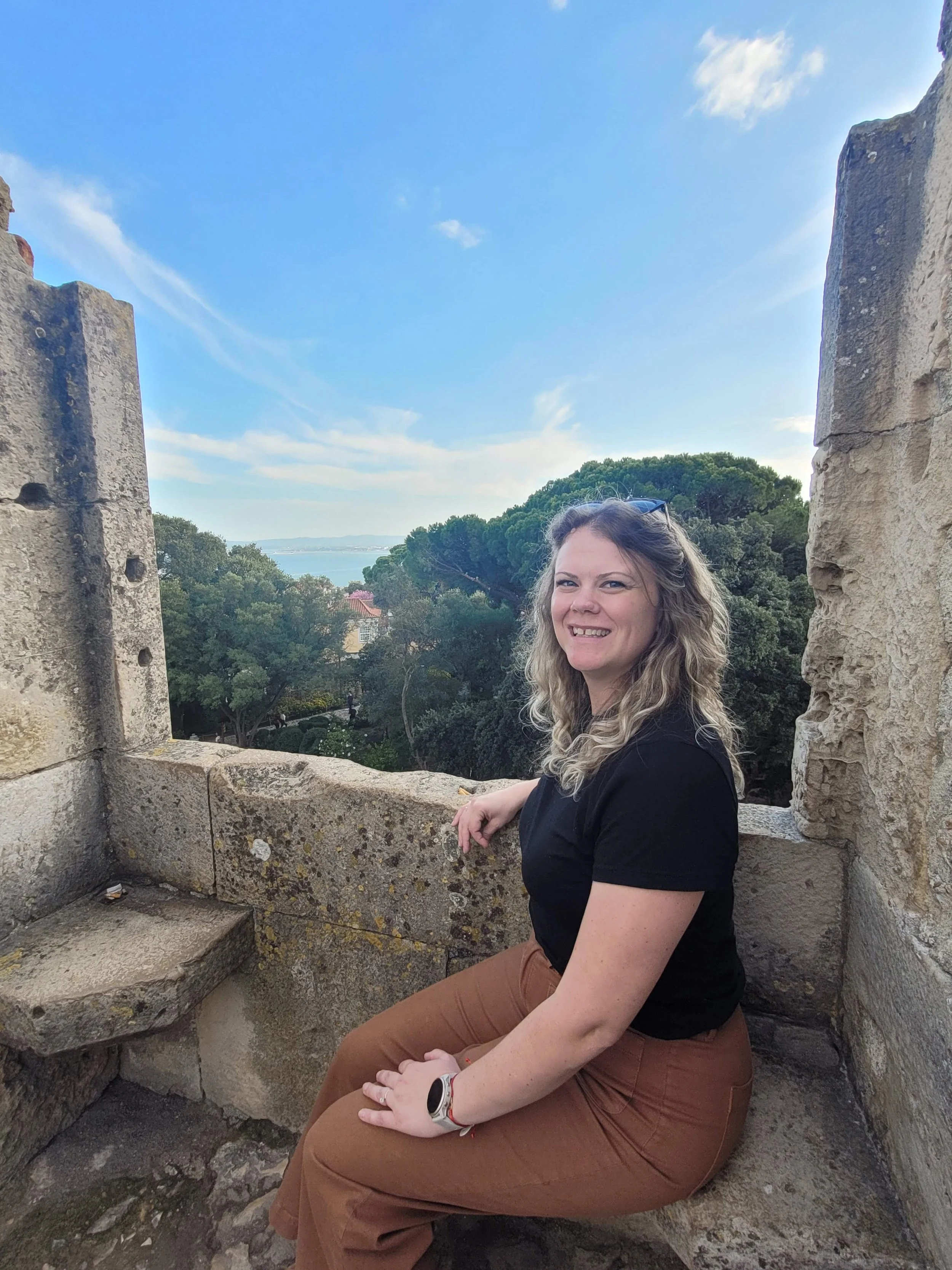 A smiling woman with curly hair, wearing a black shirt, brown pants, and a white watch, sitting on a stone structure with a scenic view of trees, water, and buildings in the background on a partly cloudy day.