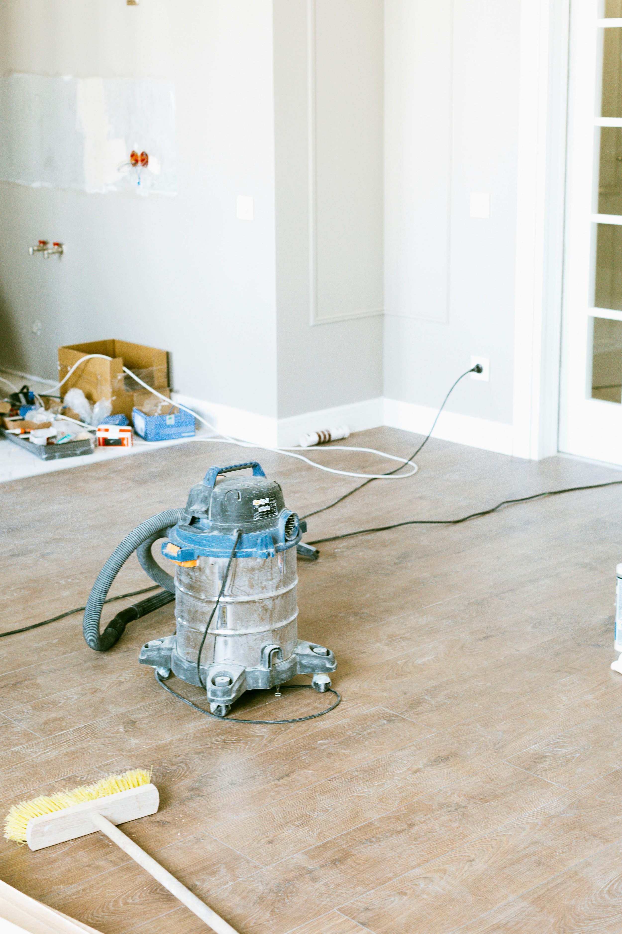 A vacuum cleaner on a wooden floor in a room under renovation, with tools and supplies scattered around, including a broom with a yellow brush and a box of various items.
