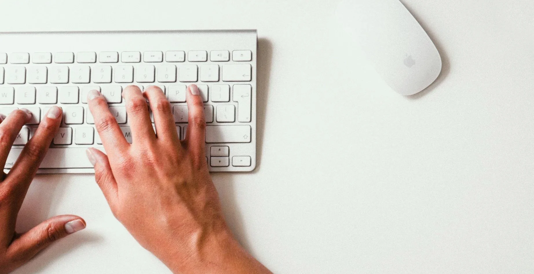 Person typing on an Apple wireless keyboard with a mouse nearby on a white surface.