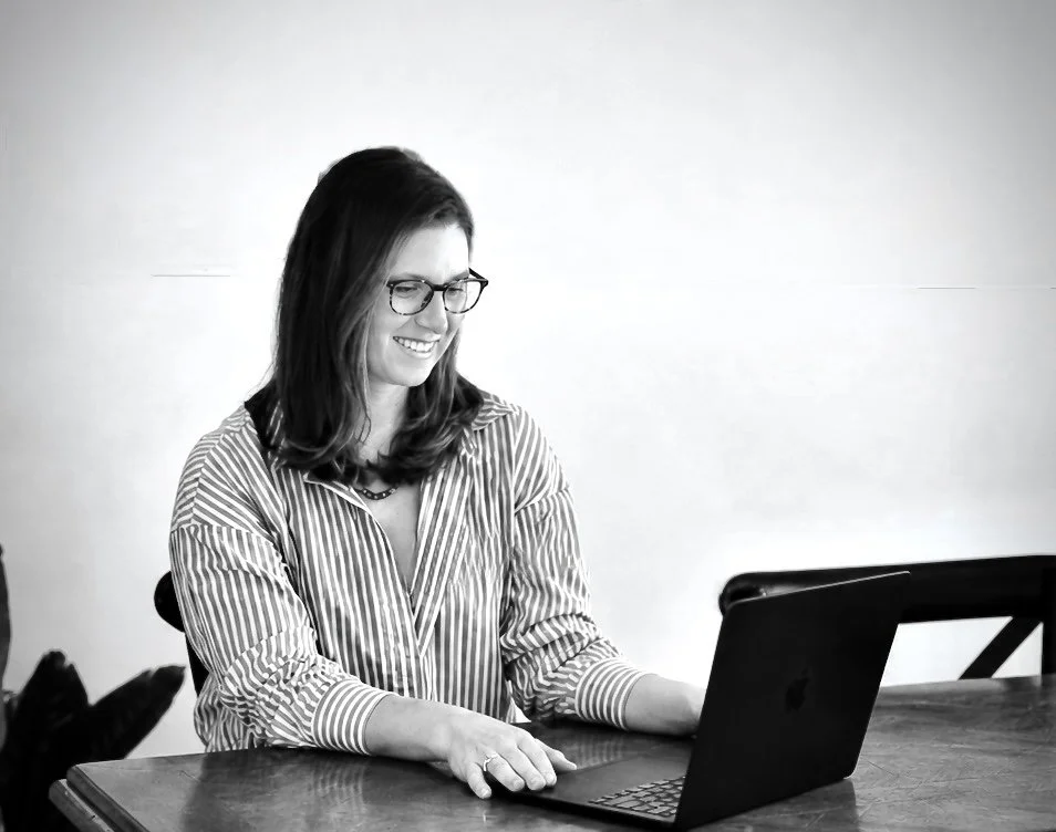 A woman with glasses and shoulder-length hair sitting at a wooden table, smiling while working on a laptop.