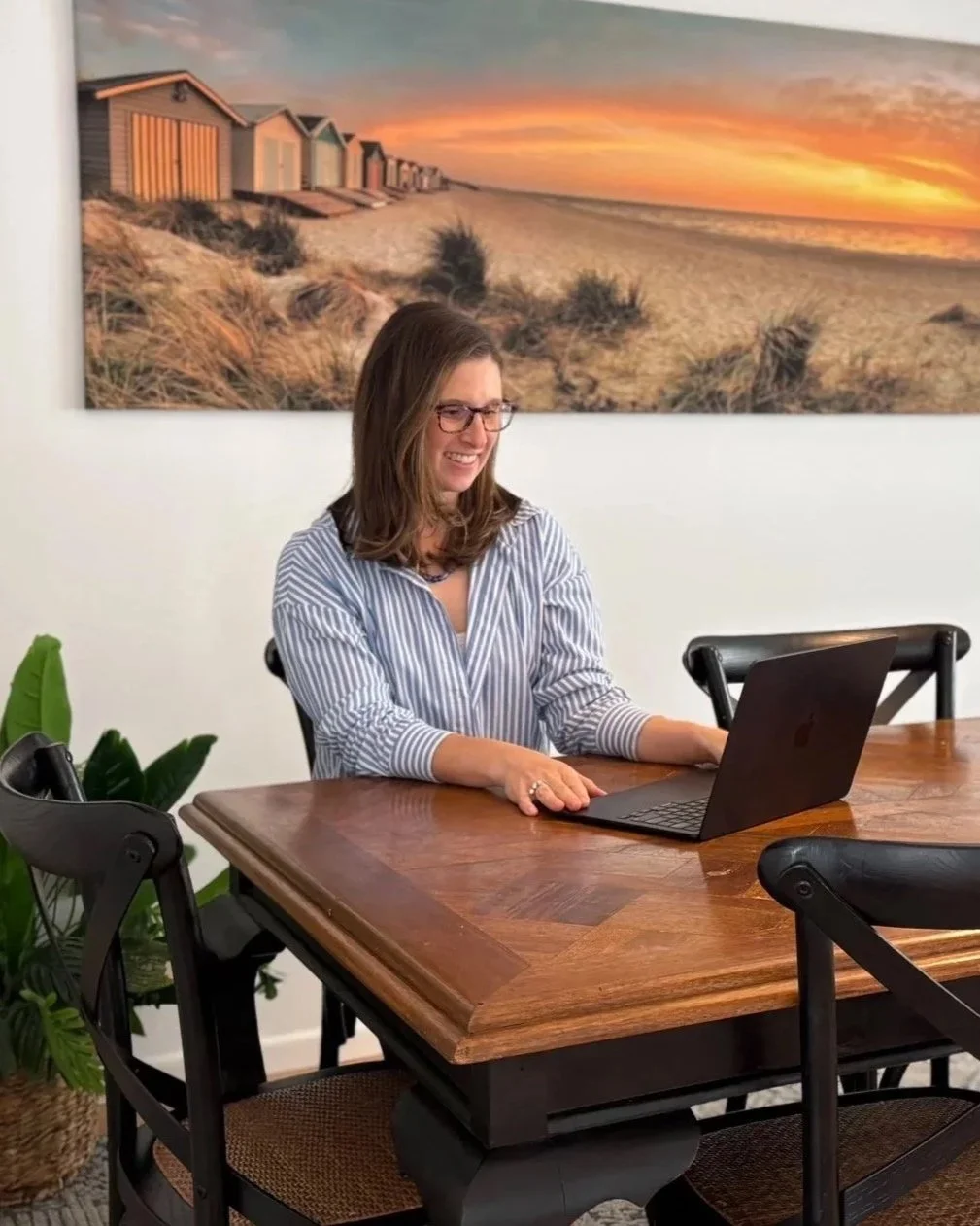 A woman sitting at a wooden table, smiling while working on a laptop. There are chairs around the table, a green plant in the left corner, and a large art photo of a beach sunset with small beach houses on the wall behind her.