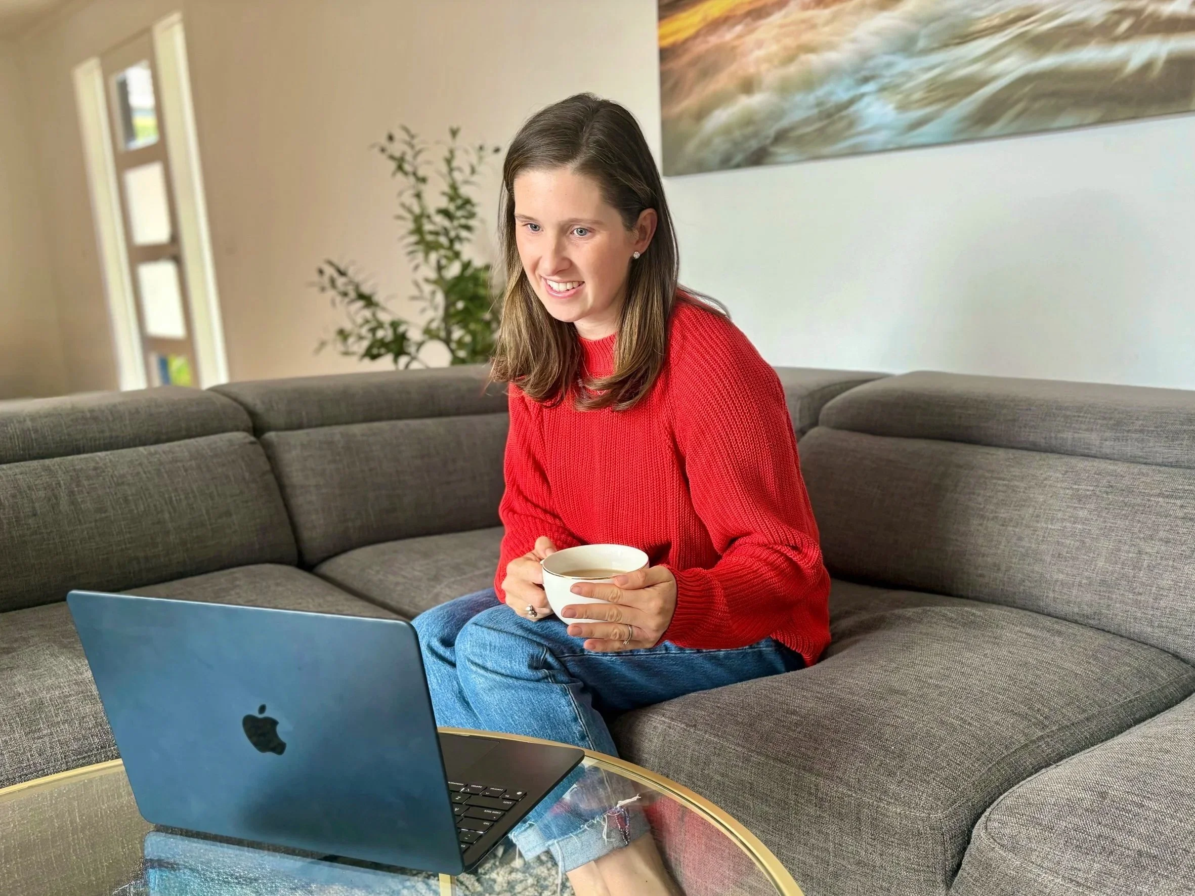 A woman with long brown hair wearing a red sweater sitting on a gray couch, holding a white cup, laptop on her lap, in a living room with a large painting on the wall behind her, a plant, and windows.