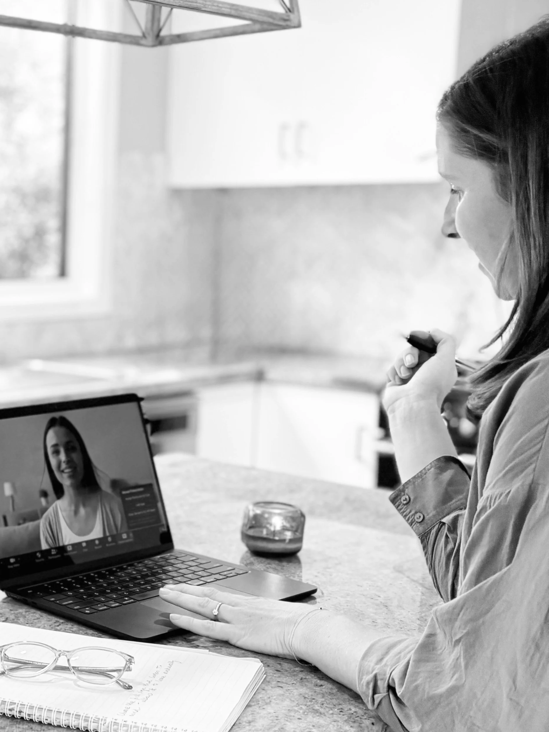 A woman appears to be working or participating in a virtual meeting during a video call on her laptop. She is sitting at a table with a notebook, a pen, and a pair of eyeglasses nearby, and there is a glass of drink in front of her. The woman is looking at her laptop screen, which shows a woman speaking in the video call.