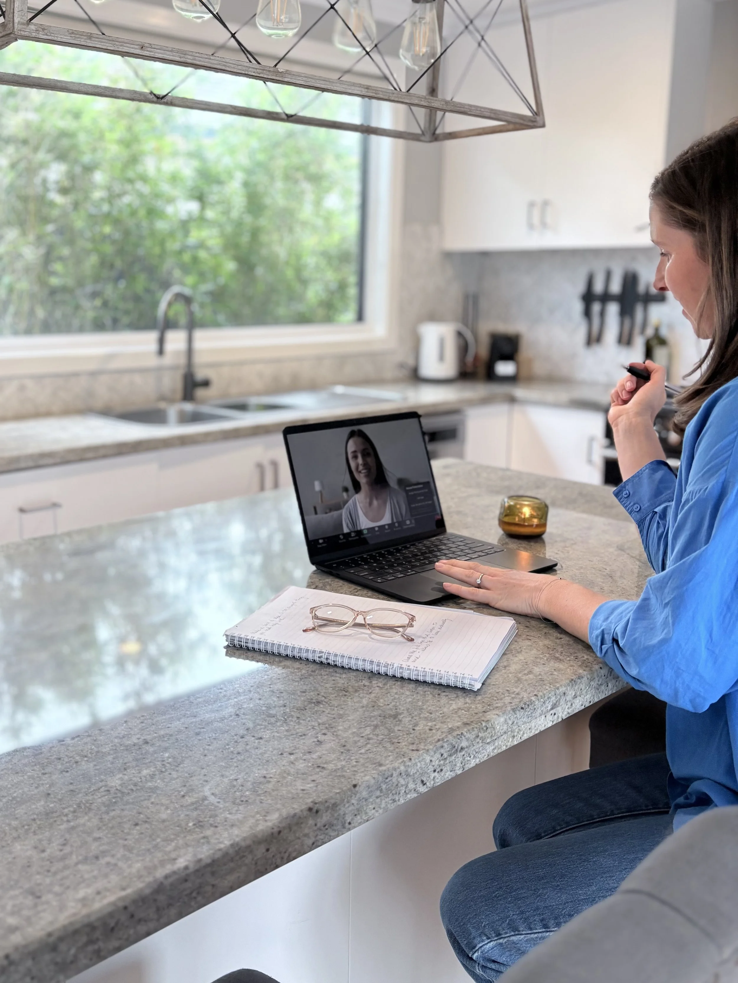 A woman is sitting at a kitchen counter, participating in a video call on her laptop. She holds a pen in one hand while looking at the screen. The counter has notebooks, glasses, and a small candle. The kitchen has a large window, white cabinets, and small appliances in the background.