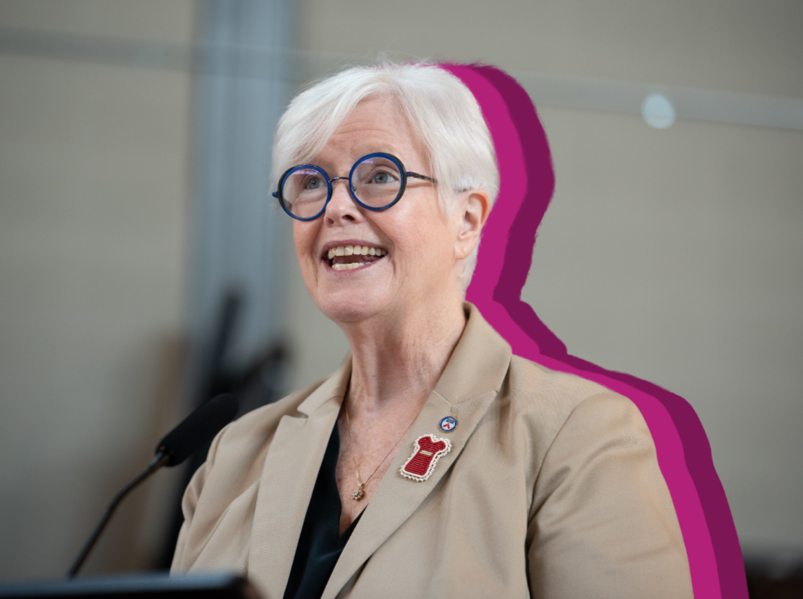 An elderly woman with short white hair, wearing glasses, a beige blazer with a red pin, and speaking at a podium.