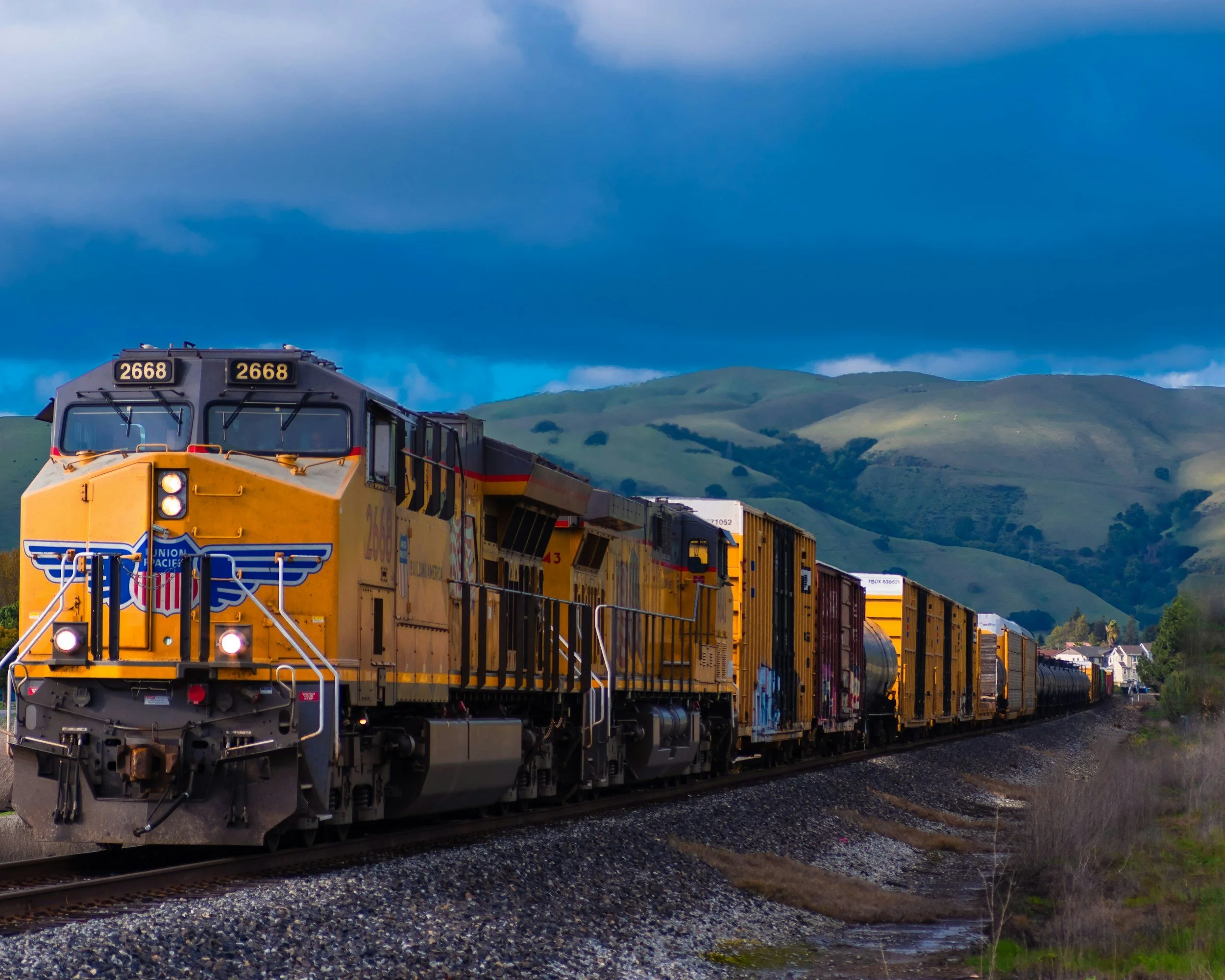 A yellow freight train moving on tracks with green hills and a cloudy sky in the background.