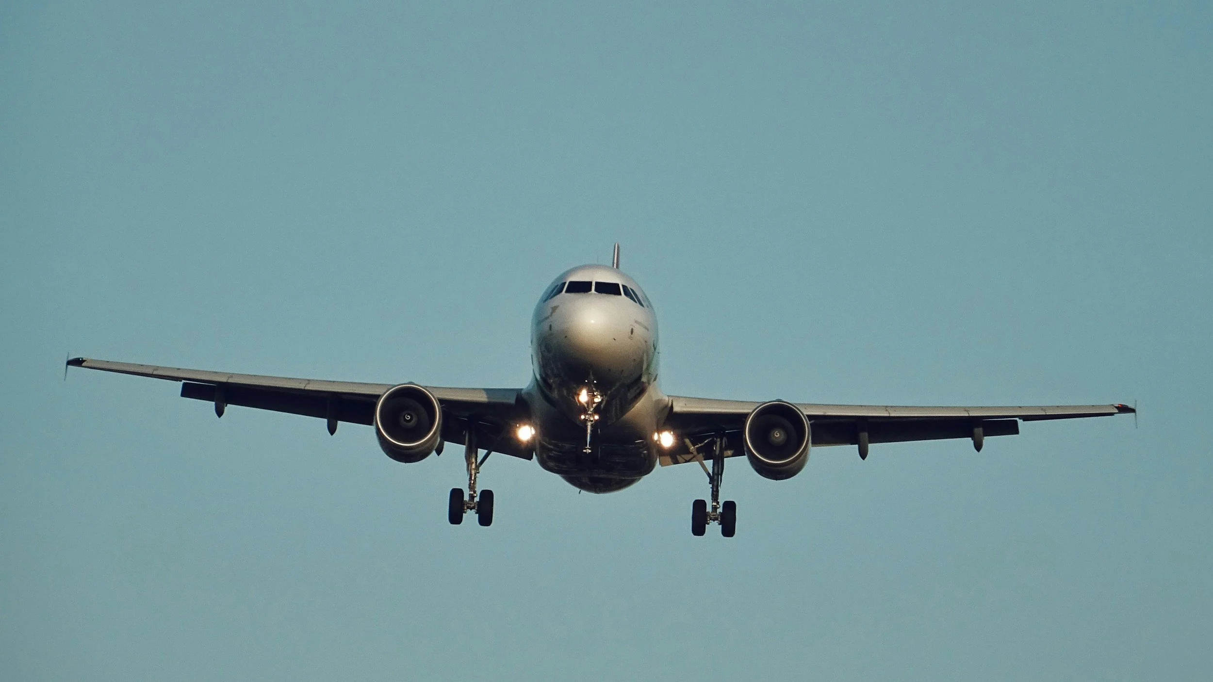 Front view of a commercial airplane descending with landing gear extended against a light blue sky.