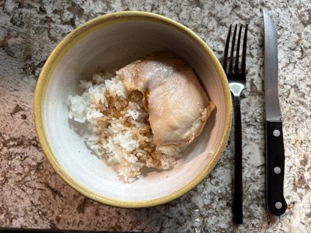 A bowl containing cooked white rice and a piece of cooked chicken, placed on a granite countertop. A fork and knife are positioned to the right of the bowl.