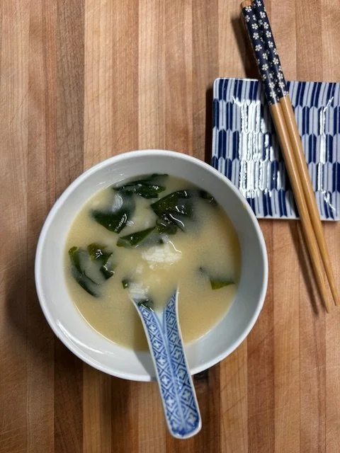 A bowl of miso soup with seaweed and tofu, a pair of chopsticks with a blue and white pattern resting on a blue and white patterned tray, on a wooden table.